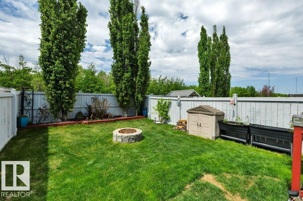 Fenced backyard featuring a stone-look fire pit, green lawn, and tall privacy trees - 6042 214 Street, Edmonton, AB - Outdoor