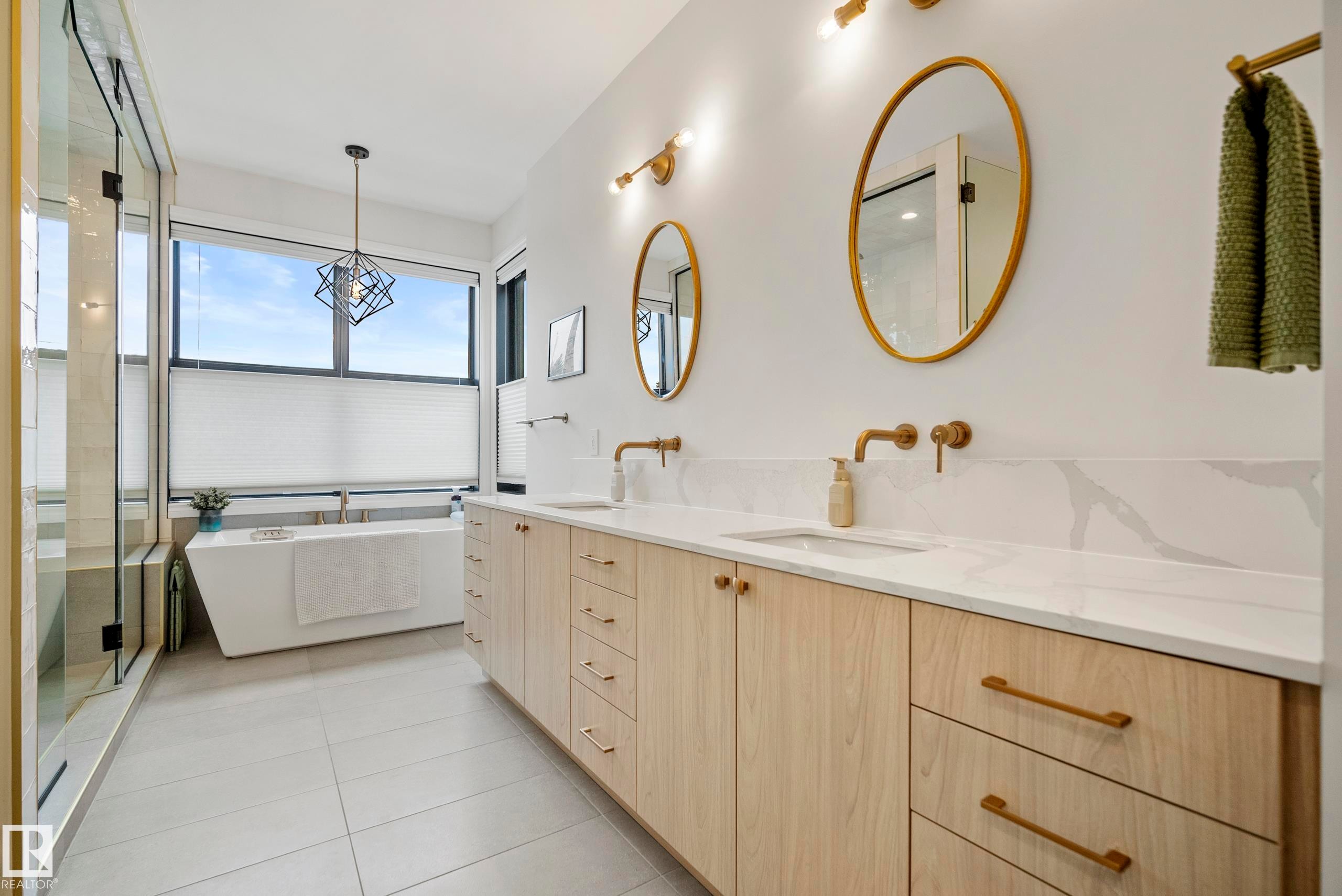Contemporary bathroom featuring a freestanding soaking tub, a spacious glass-enclosed shower, a double vanity with light wood-finish cabinetry, white stone countertops, and wall-mounted brass fixtures - 6115 111 Avenue, Edmonton, AB - Indoor Photo Showing Bathroom