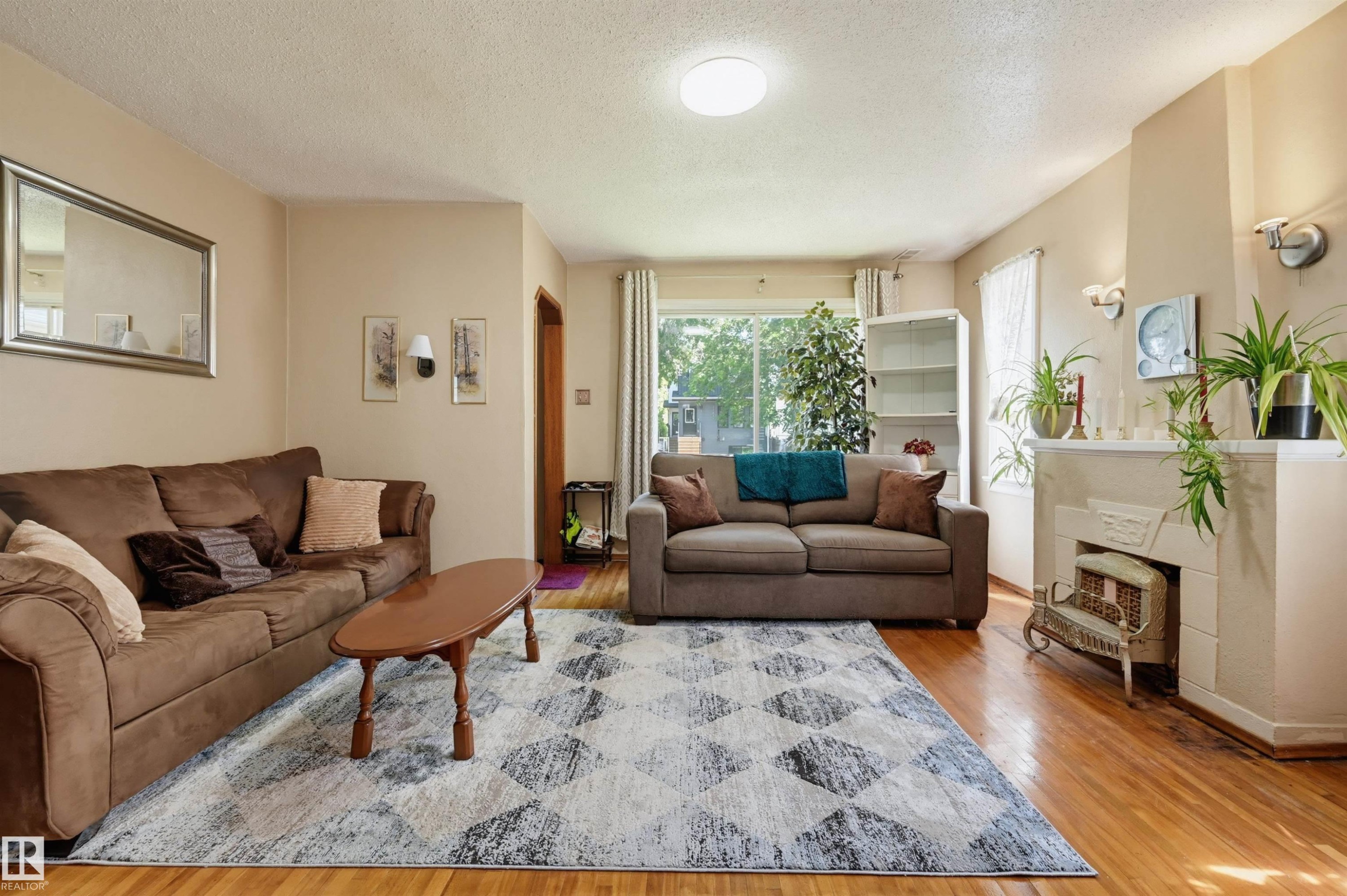 Hardwood flooring extends throughout this living space, featuring a fireplace with hearth, a large window, and wall-mounted sconces providing ambient lighting - 11414 88 Street Nw, Edmonton, AB