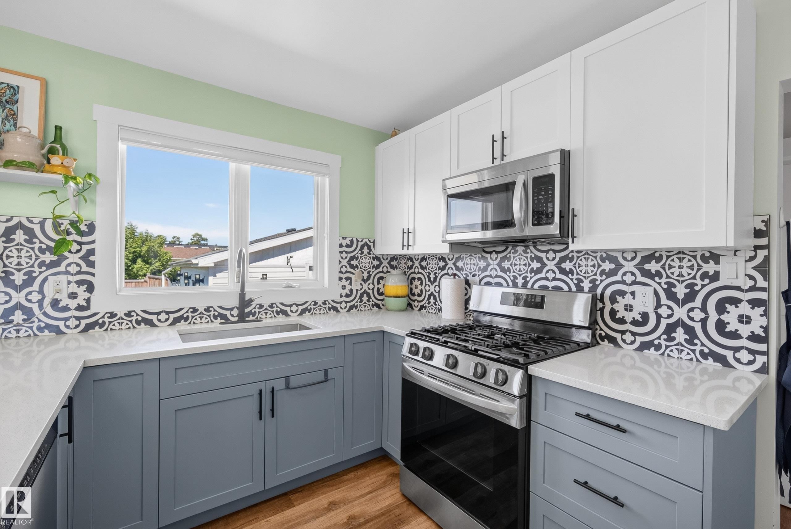 Kitchen featuring two-tone cabinetry with white upper and blue-grey lower cabinets, stainless steel appliances, patterned tile backsplash, and light-toned countertops - 14128 73 Street, Edmonton, AB