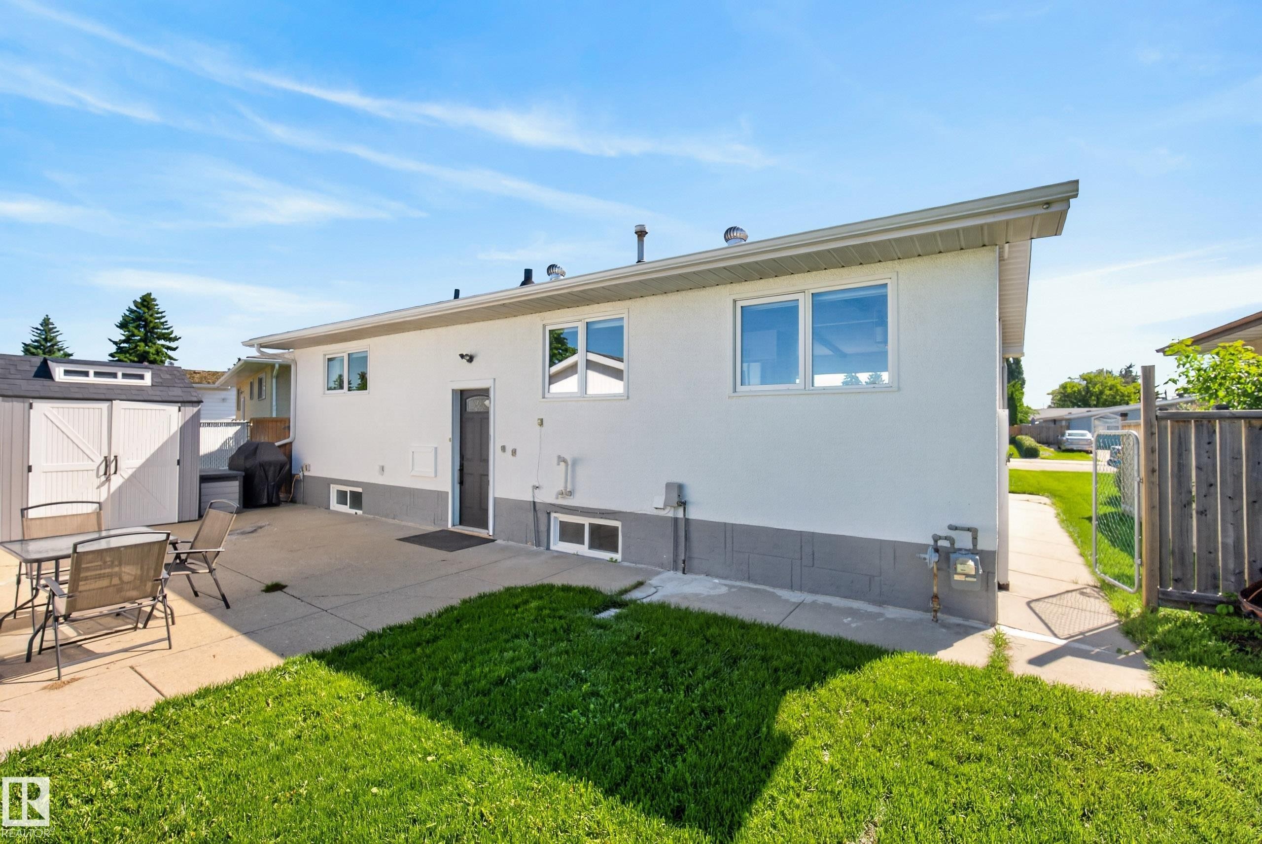 Rear exterior featuring white siding with a gray foundation, a concrete patio, and a dedicated storage shed - 14128 73 Street, Edmonton, AB