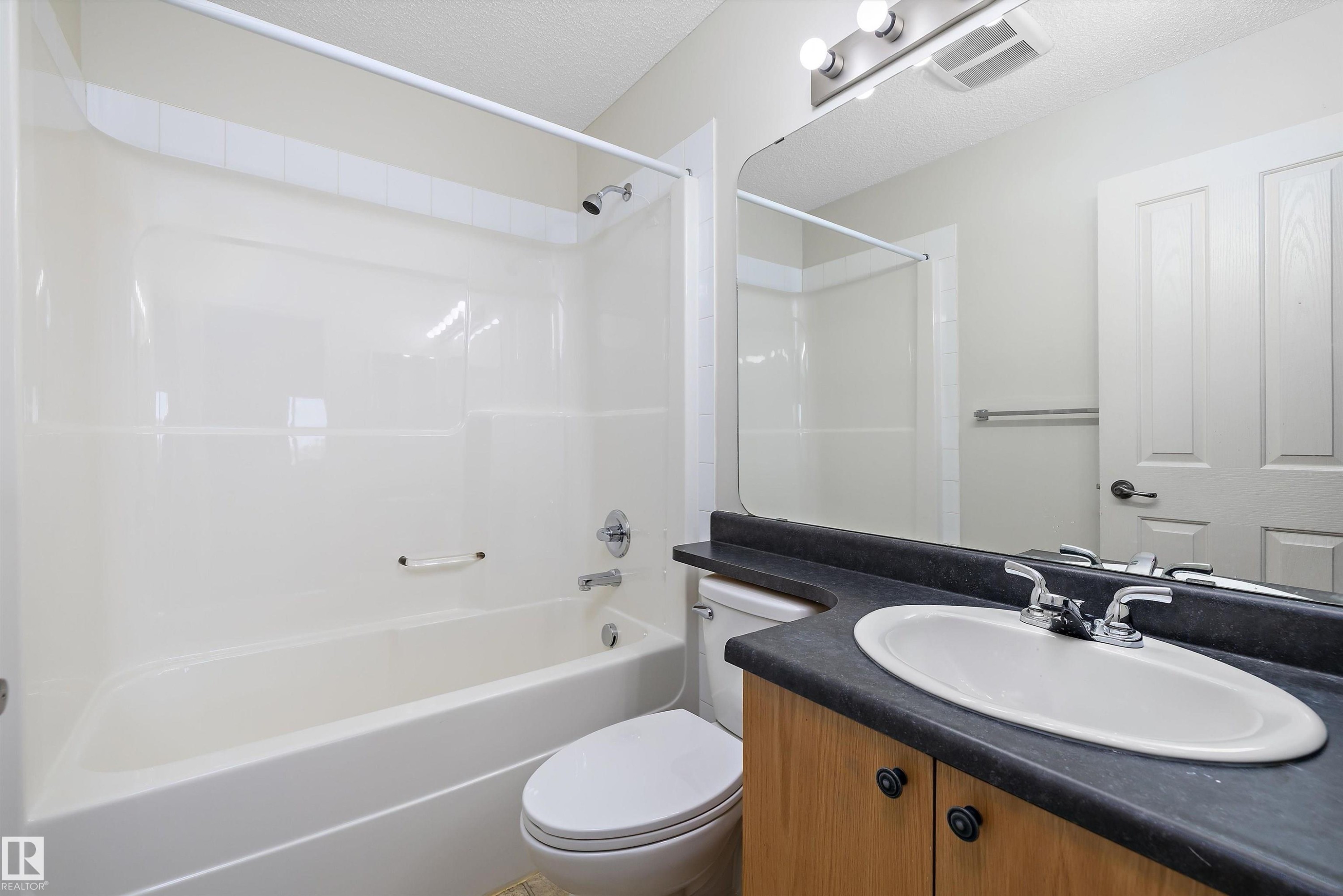 Bathroom featuring a white tub-shower combination, a vanity with a dark countertop and wood-finish cabinetry, and a mirrored wall - 101 13825 155 Avenue, Edmonton, AB