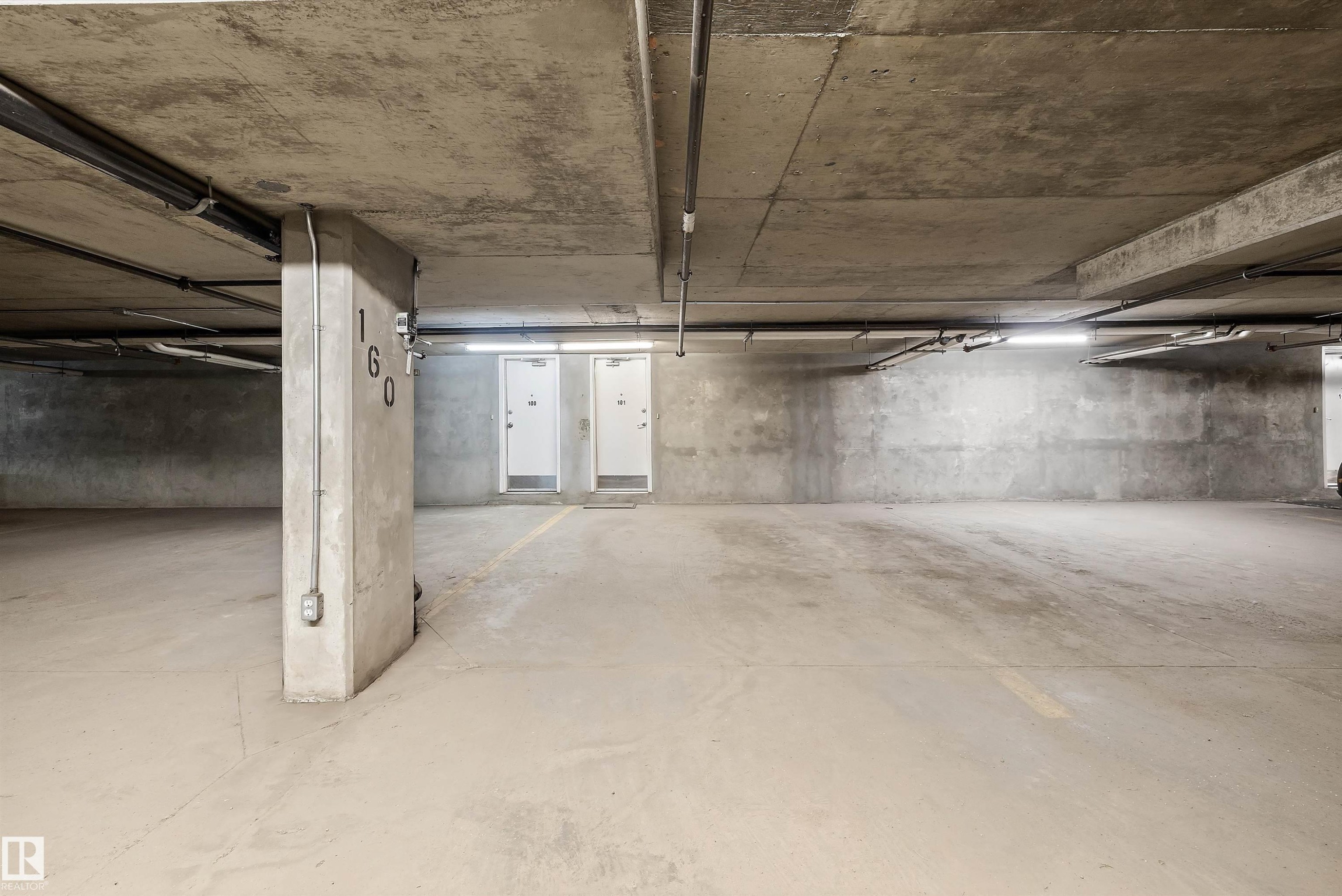 Spacious concrete parking garage featuring overhead lighting, exposed conduit, and multiple access doors - 101 13825 155 Avenue, Edmonton, AB