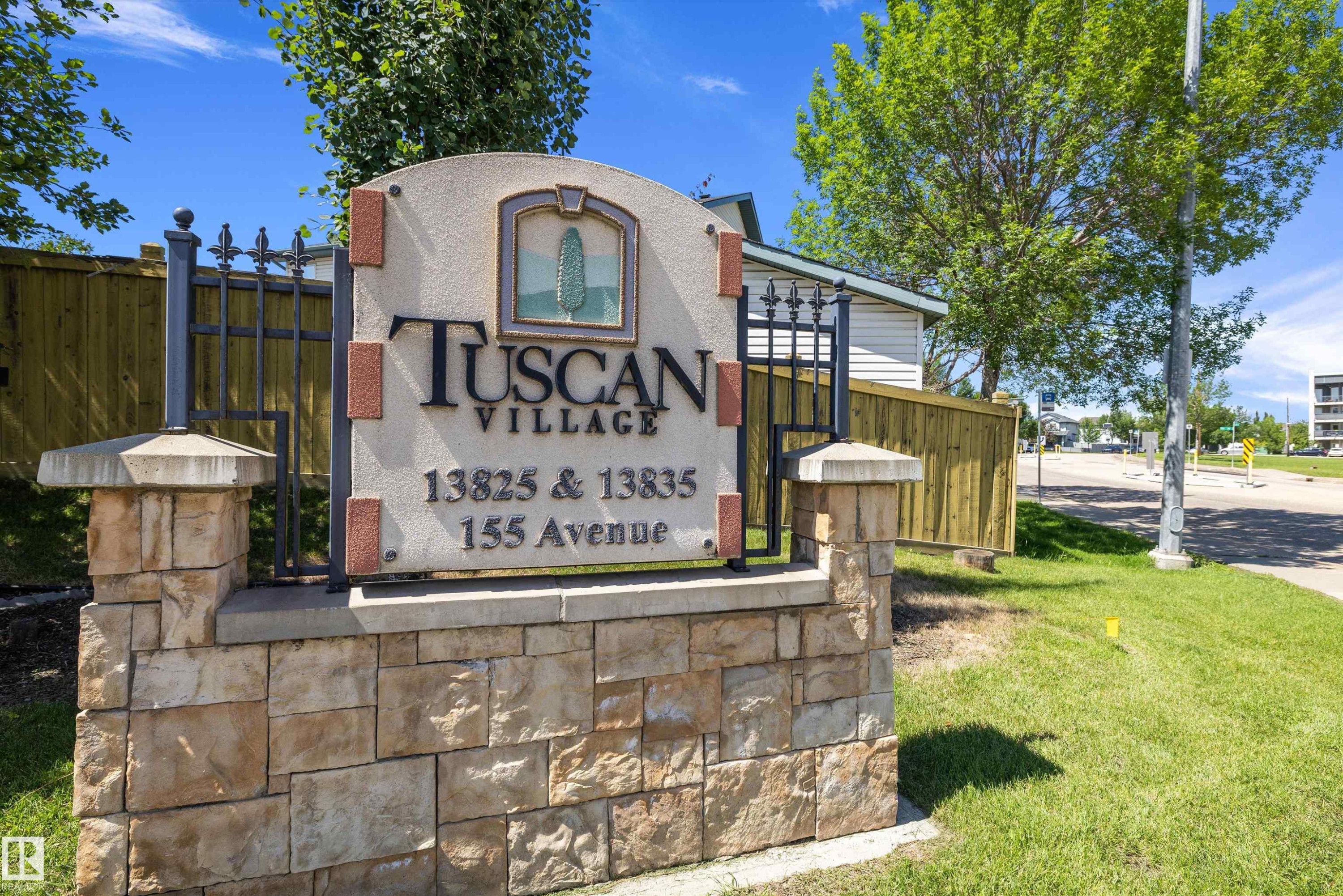 "Tuscan Village" entrance sign featuring a stone base, wrought iron detailing, and an arched stucco panel with a decorative emblem - 101 13825 155 Avenue, Edmonton, AB