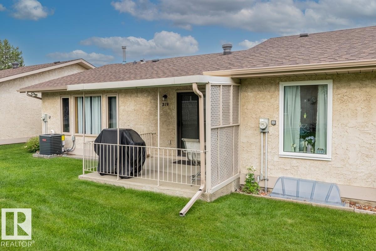 Exterior featuring stucco siding, a covered patio with railing, and a window well cover - 219 Knottwood Road, Edmonton, AB