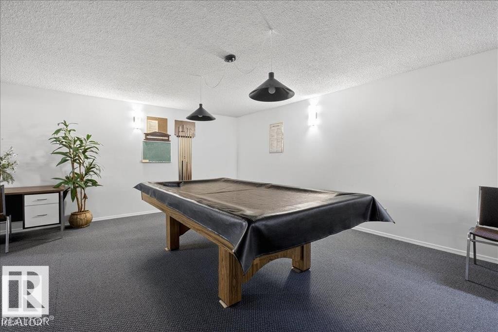 Spacious recreation room featuring a wooden billiard table, textured ceiling, and dark carpet flooring - 219 Knottwood Road, Edmonton, AB