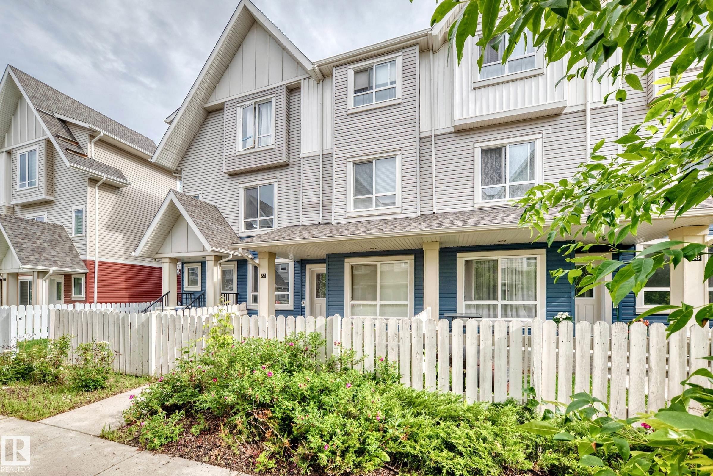 Modern townhome exterior featuring multi-level construction, durable siding, a pitched roofline, white picket fencing, and mature landscaping - 37 13003 132 Avenue Nw, Edmonton, AB