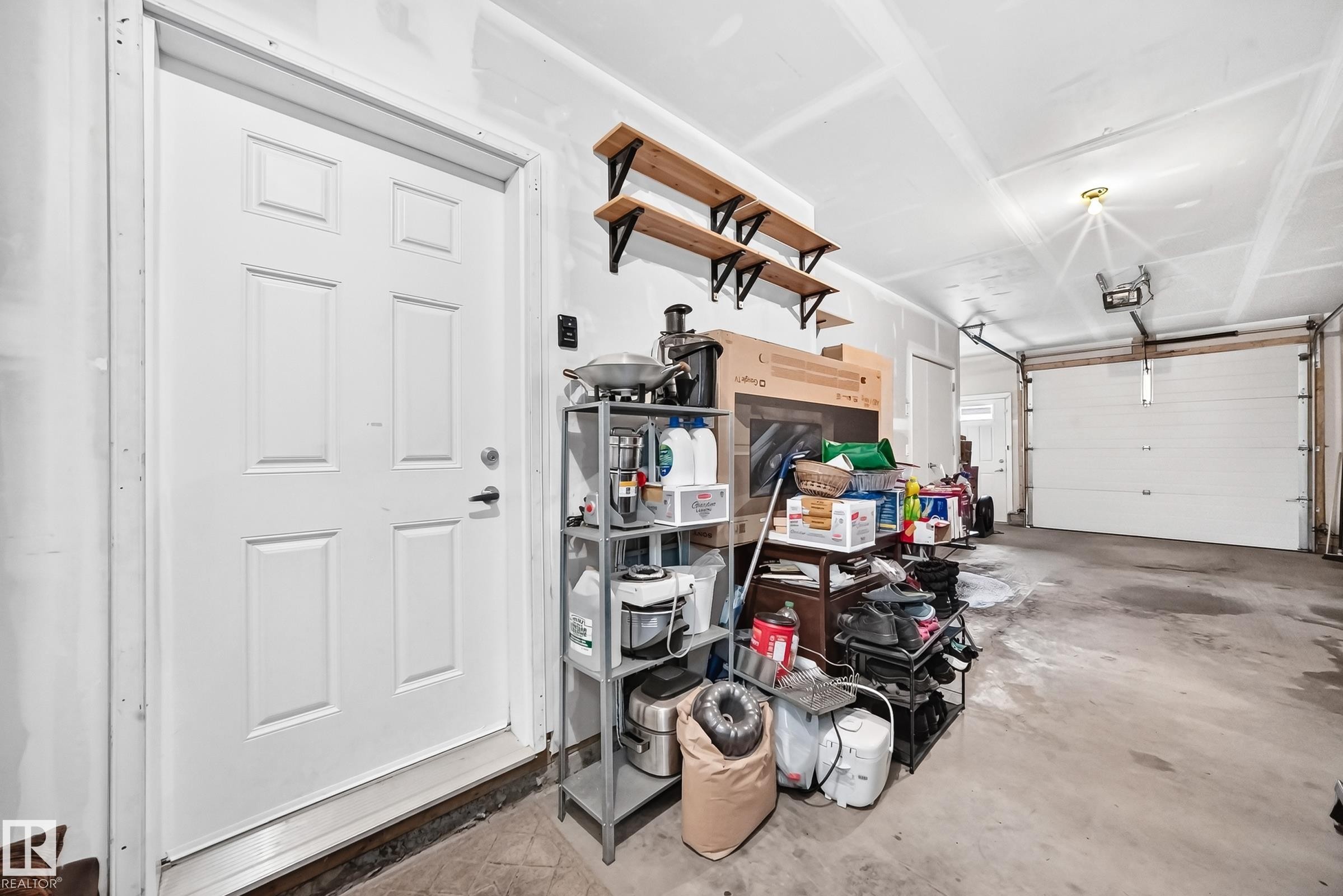 Concrete floor garage with a paneled white door, overhead garage door, built-in wall shelving, and exposed ceiling joists - 37 13003 132 Avenue Nw, Edmonton, AB