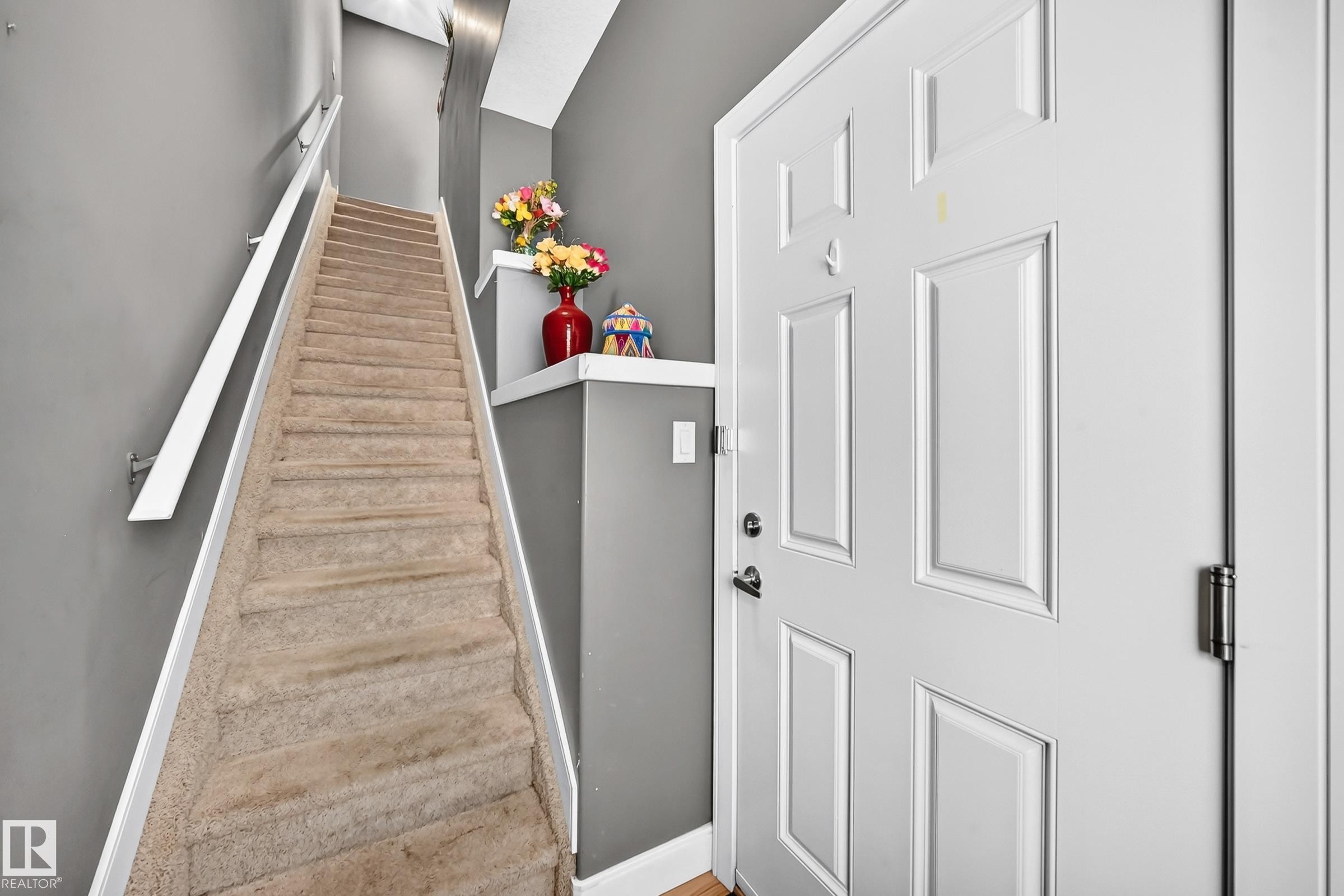 Carpeted staircase with white handrail and trim, alongside a paneled entrance door with dark hardware - 37 13003 132 Avenue Nw, Edmonton, AB