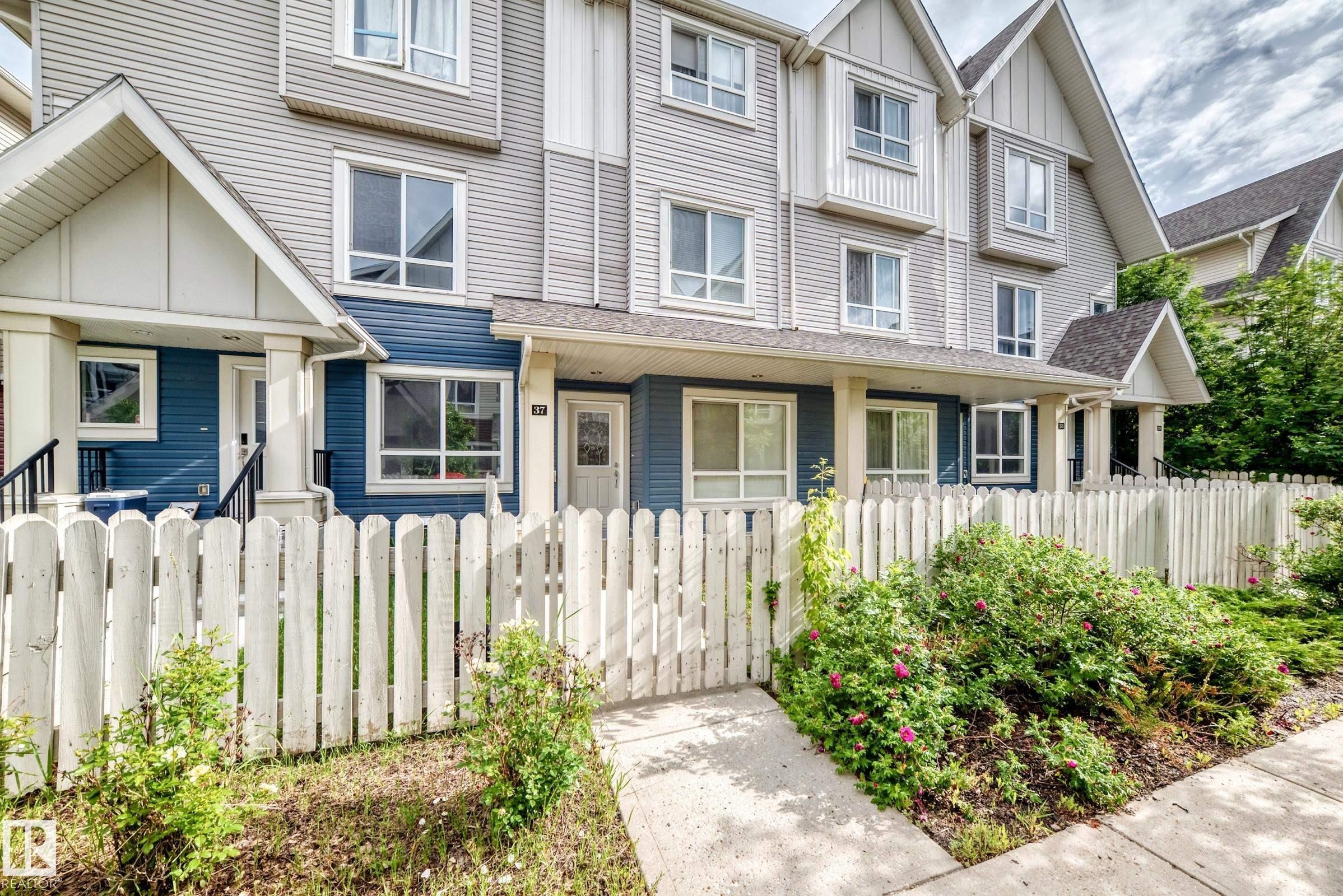 Townhome exterior featuring a white picket fence, a mature landscaped garden, blue and grey siding, multiple windows, and a covered front porch - 37 13003 132 Avenue Nw, Edmonton, AB