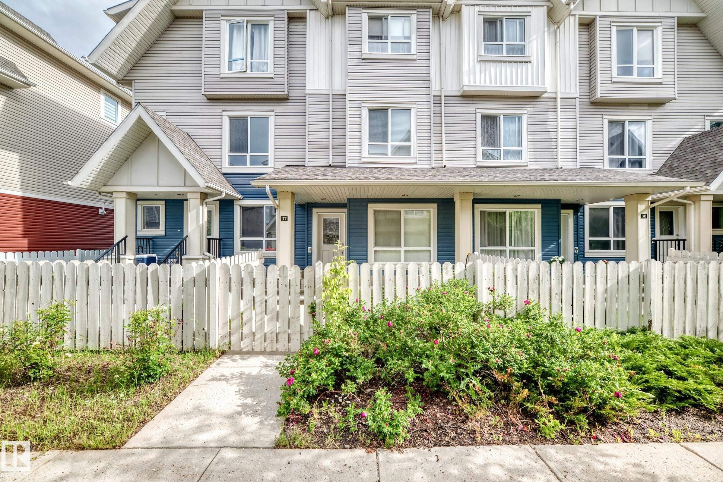 Townhome exterior featuring blue and gray siding, multiple windows, and a white picket fence - 37 13003 132 Avenue Nw, Edmonton, AB