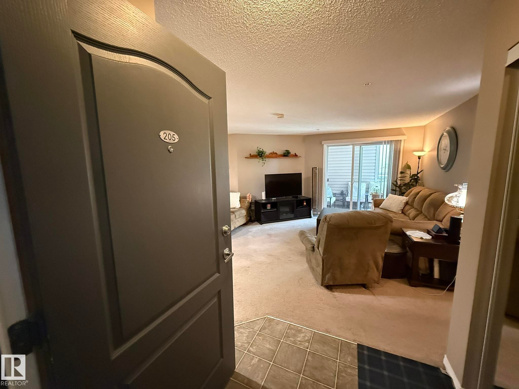 Entryway with tile flooring opening into a carpeted living space featuring a sliding glass door and a wall-mounted shelf - 205 3425 19 Street, Edmonton, AB