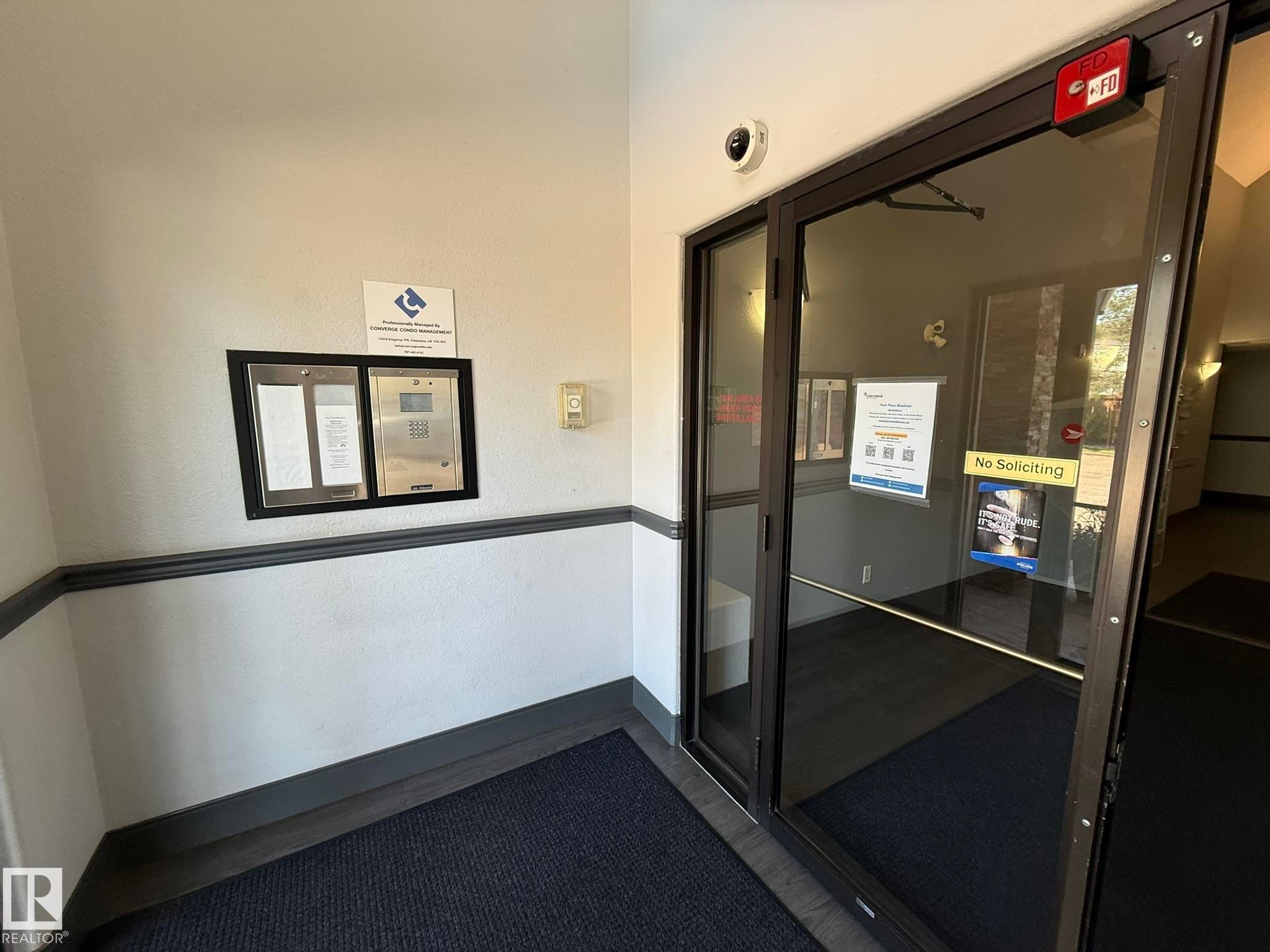 Entryway featuring a dark framed glass door, wall-mounted intercom system, and a security camera - 205 3425 19 Street, Edmonton, AB