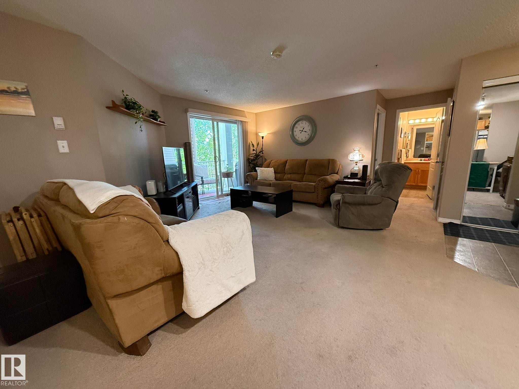Living area featuring light-toned carpeting, neutral wall paint, and a sliding glass door providing access to an outdoor balcony - 205 3425 19 Street, Edmonton, AB