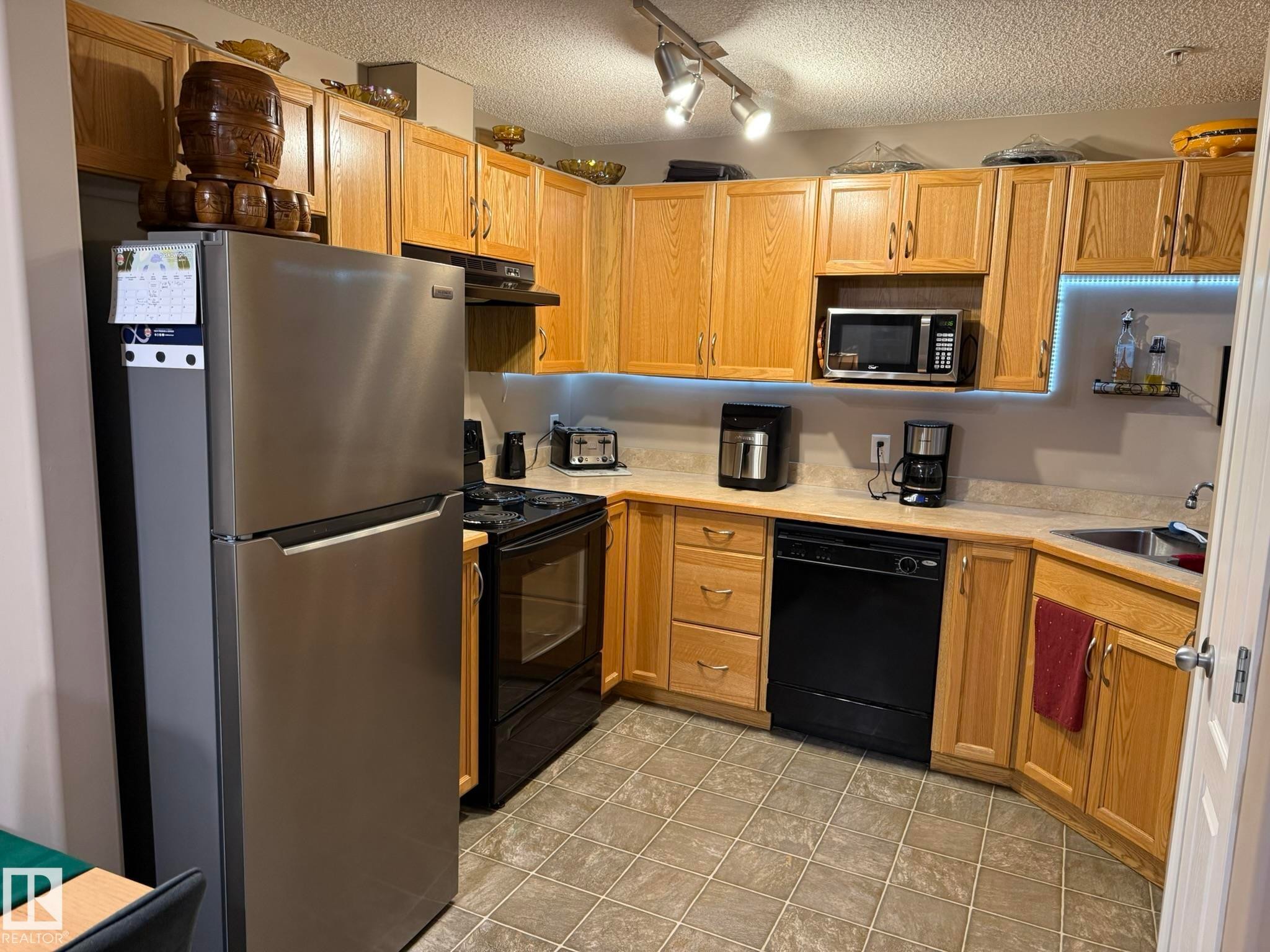 U-shaped kitchen featuring wood-finish cabinetry, a stainless steel refrigerator, black range, black dishwasher, and a stainless steel sink - 205 3425 19 Street, Edmonton, AB