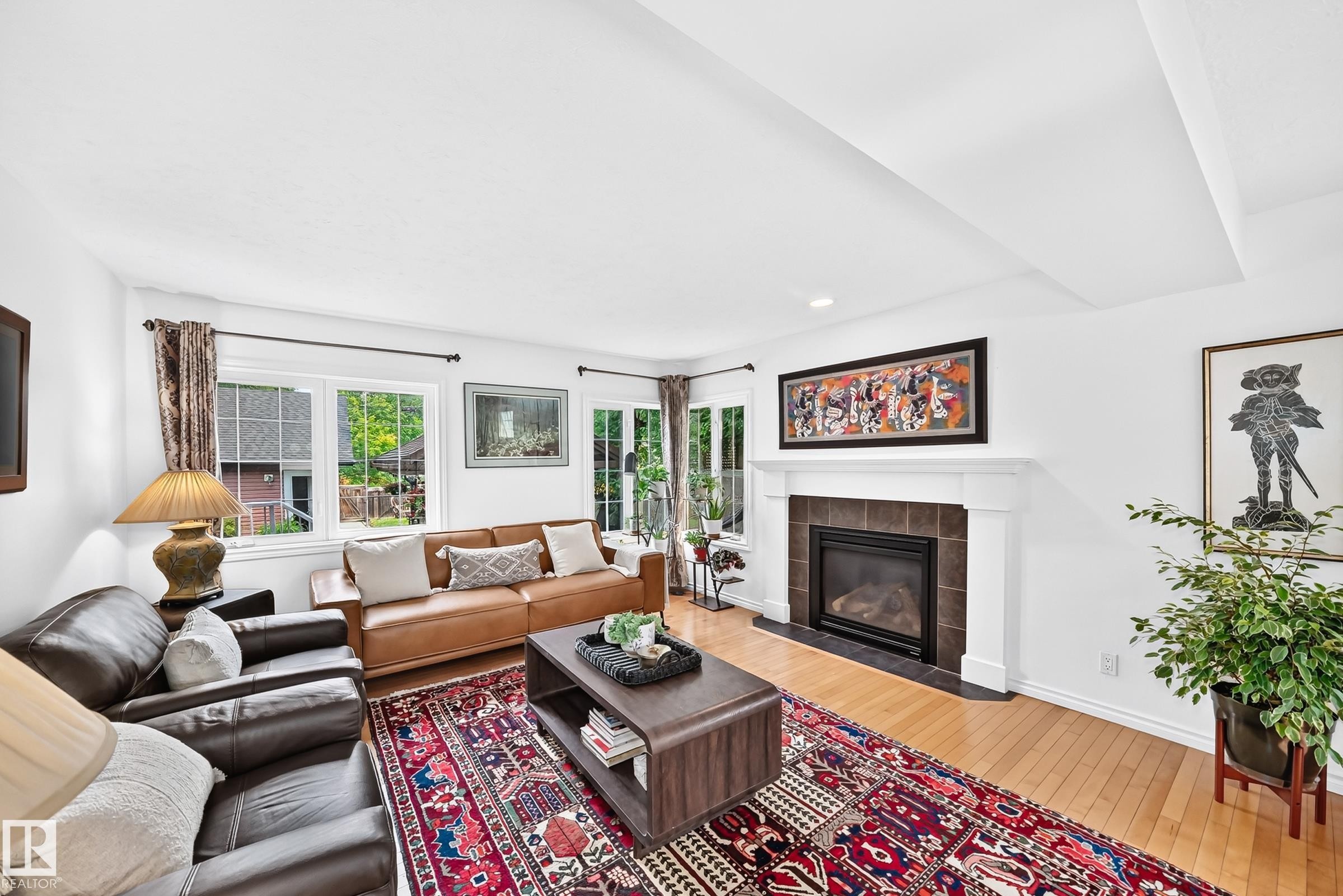 Living area featuring a fireplace with a white mantel and dark tile surround, light wood-finish flooring, and multiple windows - 10323 138 Street Nw, Edmonton, AB - Indoor Photo Showing Living Room With Fireplace
