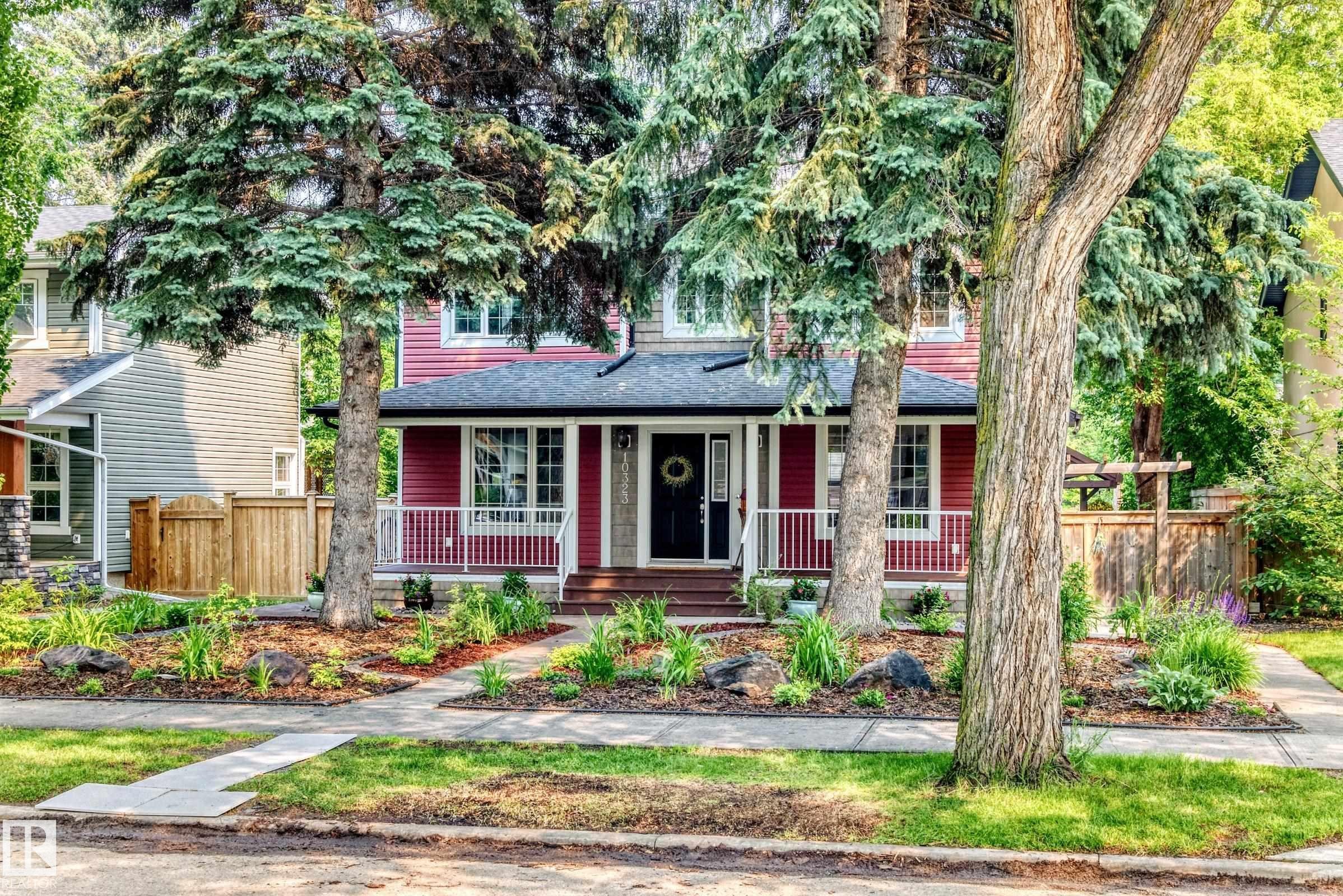 Red siding exterior with white trim, featuring a covered front porch with white railings, a dark entry door, and multiple mature trees - 10323 138 Street Nw, Edmonton, AB - Outdoor With Deck Patio Veranda