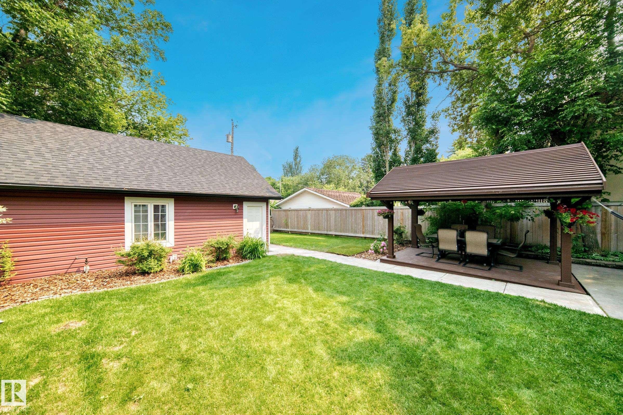 Expansive backyard featuring a large lawn area, a side building with red siding and a shingled roof, and a covered outdoor patio with a composite deck base - 10323 138 Street Nw, Edmonton, AB - Outdoor