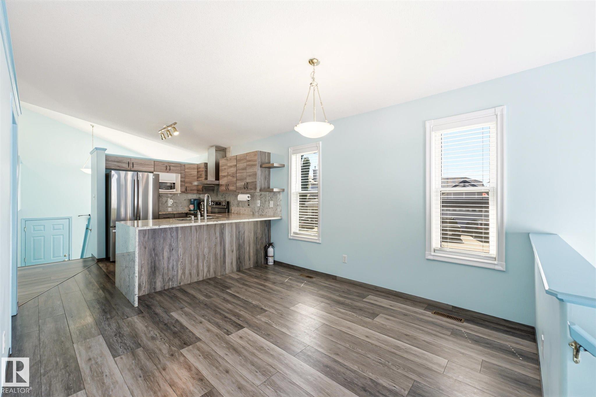 Open-concept kitchen and dining area featuring wood-finish flooring, a kitchen island with stone countertop, stainless steel appliances, and two windows with blinds - 14908 137 Street Nw, Edmonton, AB - Indoor Photo Showing Kitchen