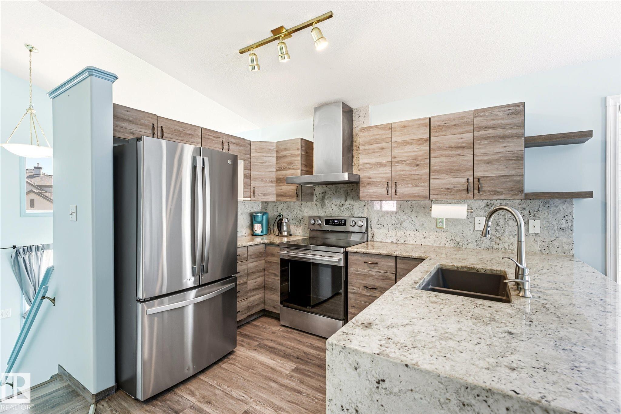 Contemporary kitchen featuring a large stainless steel French door refrigerator, wood-finish cabinetry, light-toned countertops and backsplash, stainless steel range with overhead hood, and a deep basin sink with a high-arc faucet - 14908 137 Street Nw, Edmonton, AB - Indoor Photo Showing Kitchen With Stainless Steel Kitchen With Double Sink With Upgraded Kitchen