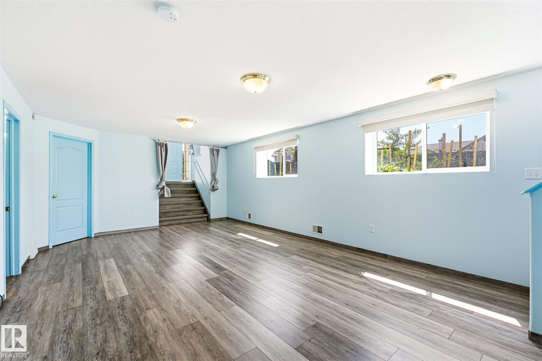 Spacious room featuring wood-finish flooring, two windows with roller shades, and light blue interior doors - 14908 137 Street Nw, Edmonton, AB - Indoor