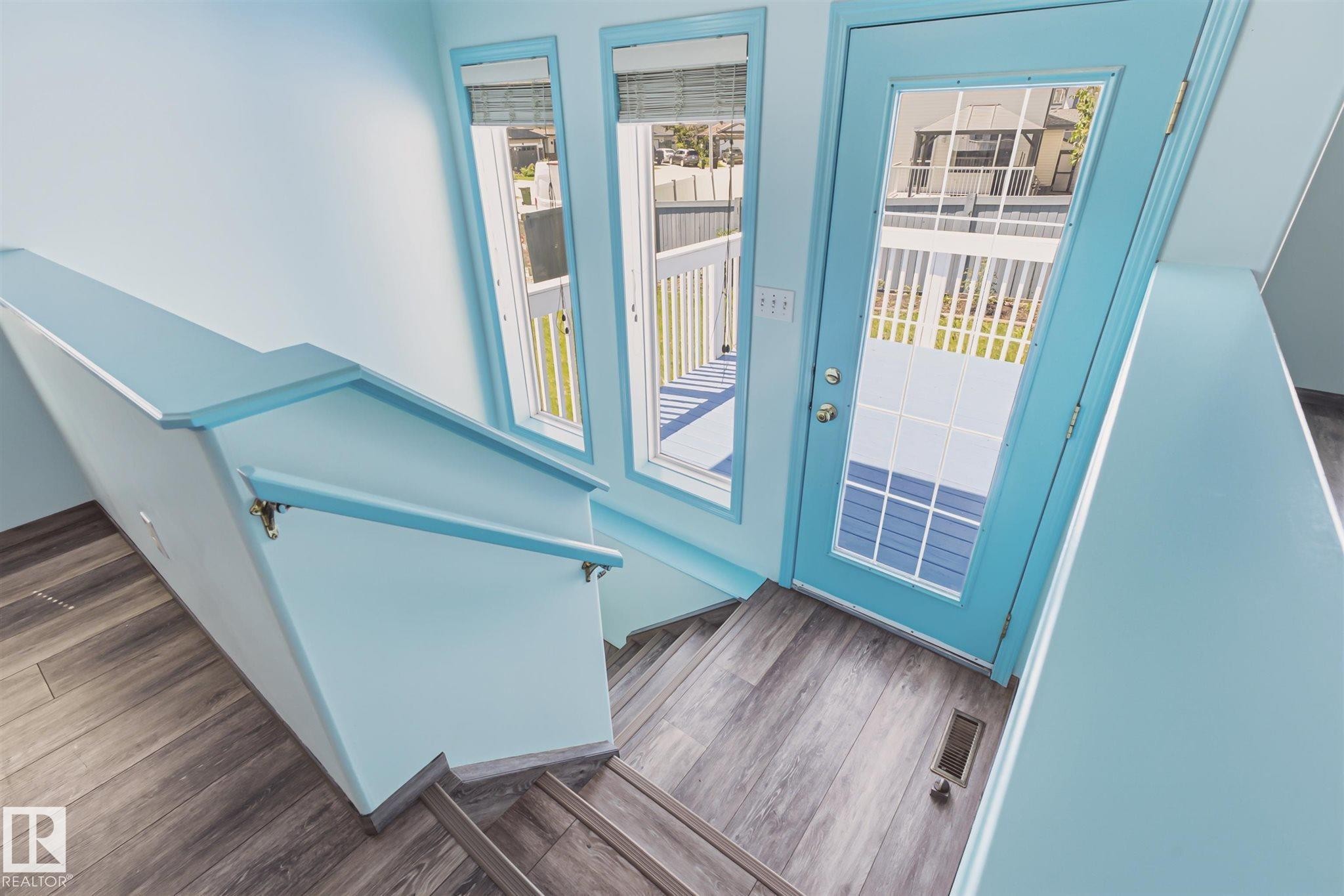 Stairwell featuring wood-finish flooring, a light blue handrail, three windows, and a glass-paneled door opening to a deck - 14908 137 Street Nw, Edmonton, AB - Indoor Photo Showing Other Room