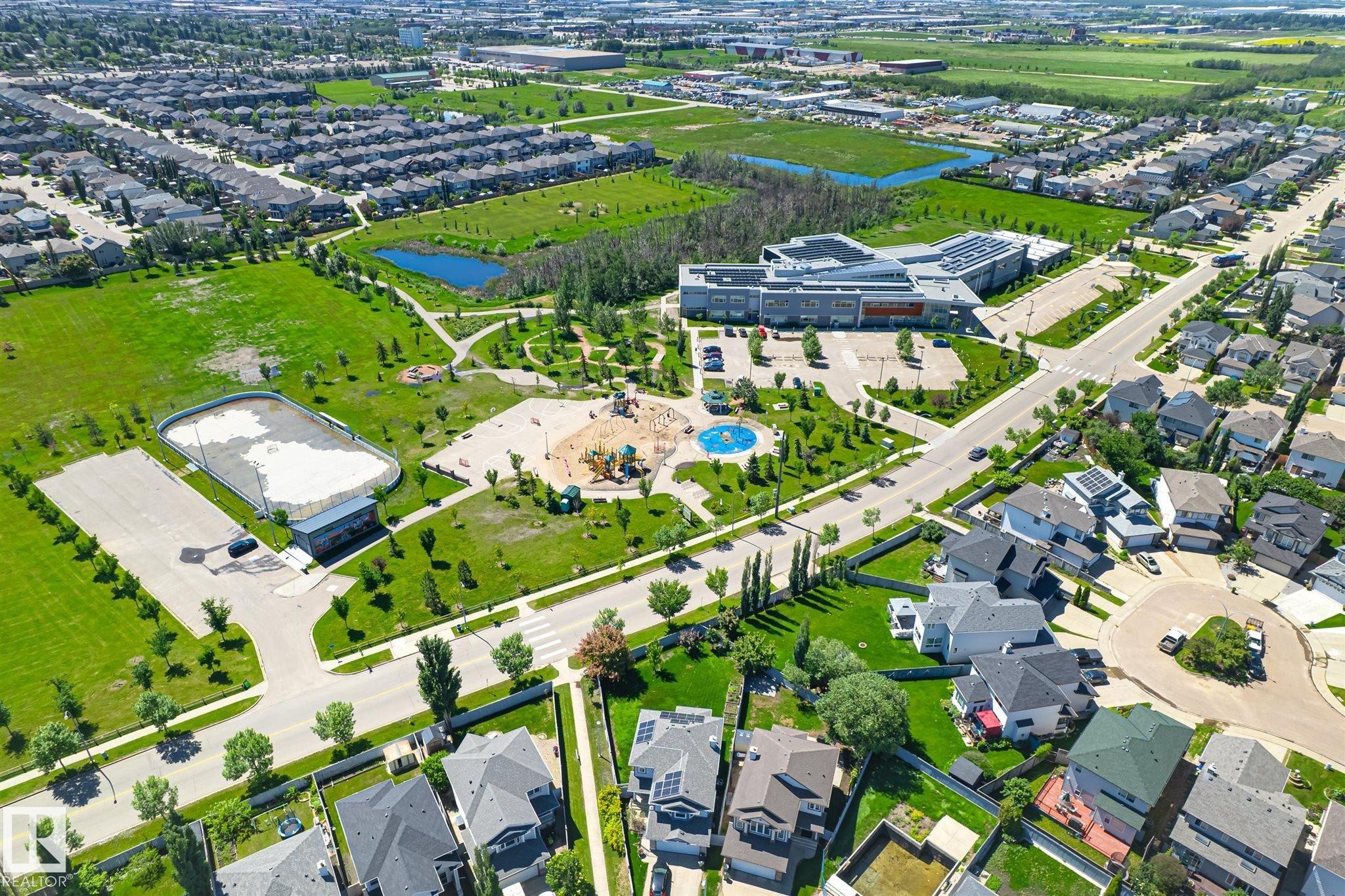 Aerial view of a residential community featuring extensive green spaces, a large recreational park with playground equipment, a modern institutional building, and a distinct sports court - 14908 137 Street Nw, Edmonton, AB - Outdoor With View