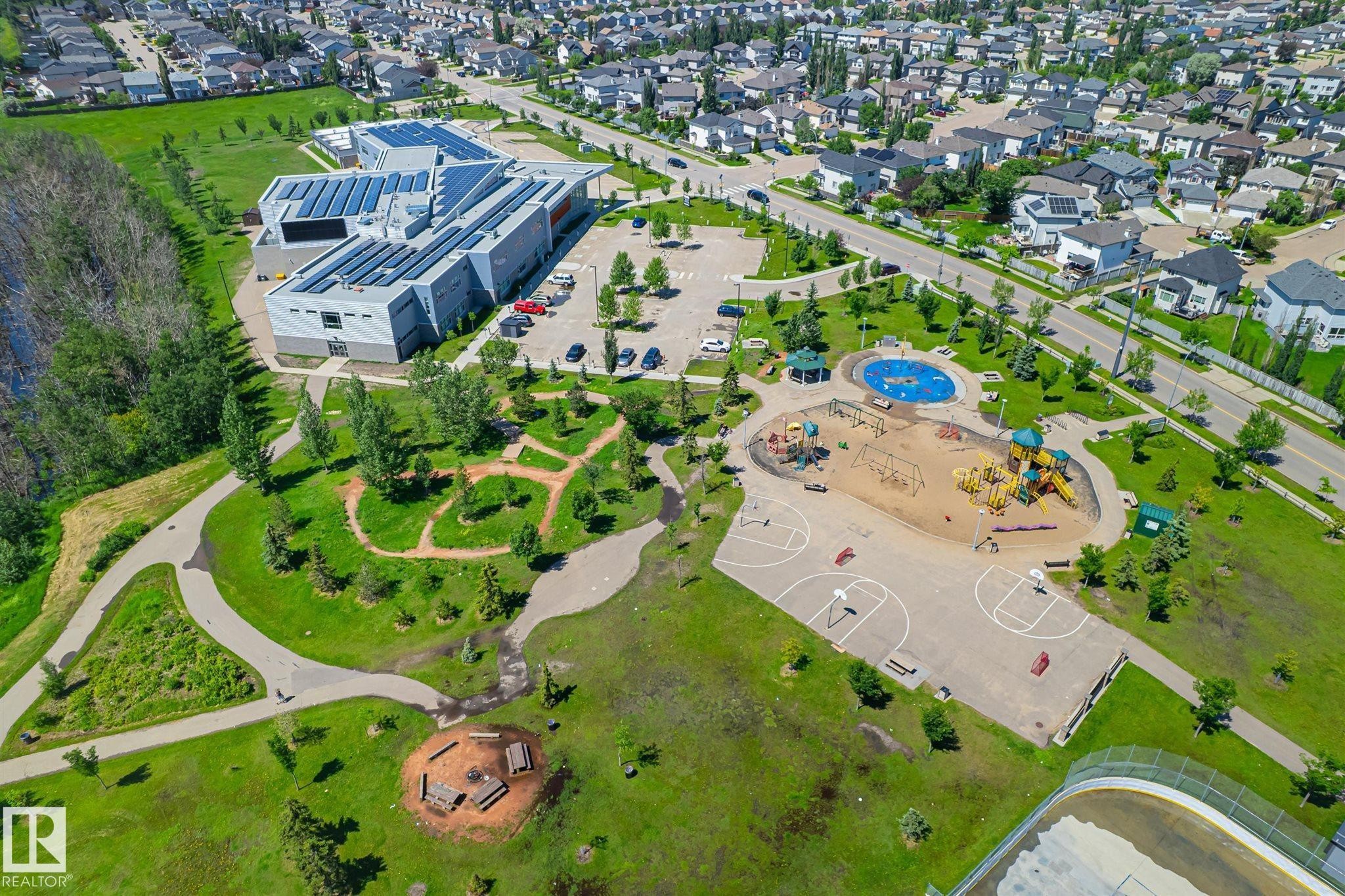 Modern community building with extensive rooftop solar panels, surrounded by green spaces, paved walking paths, and a public playground area - 14908 137 Street Nw, Edmonton, AB - Outdoor With View
