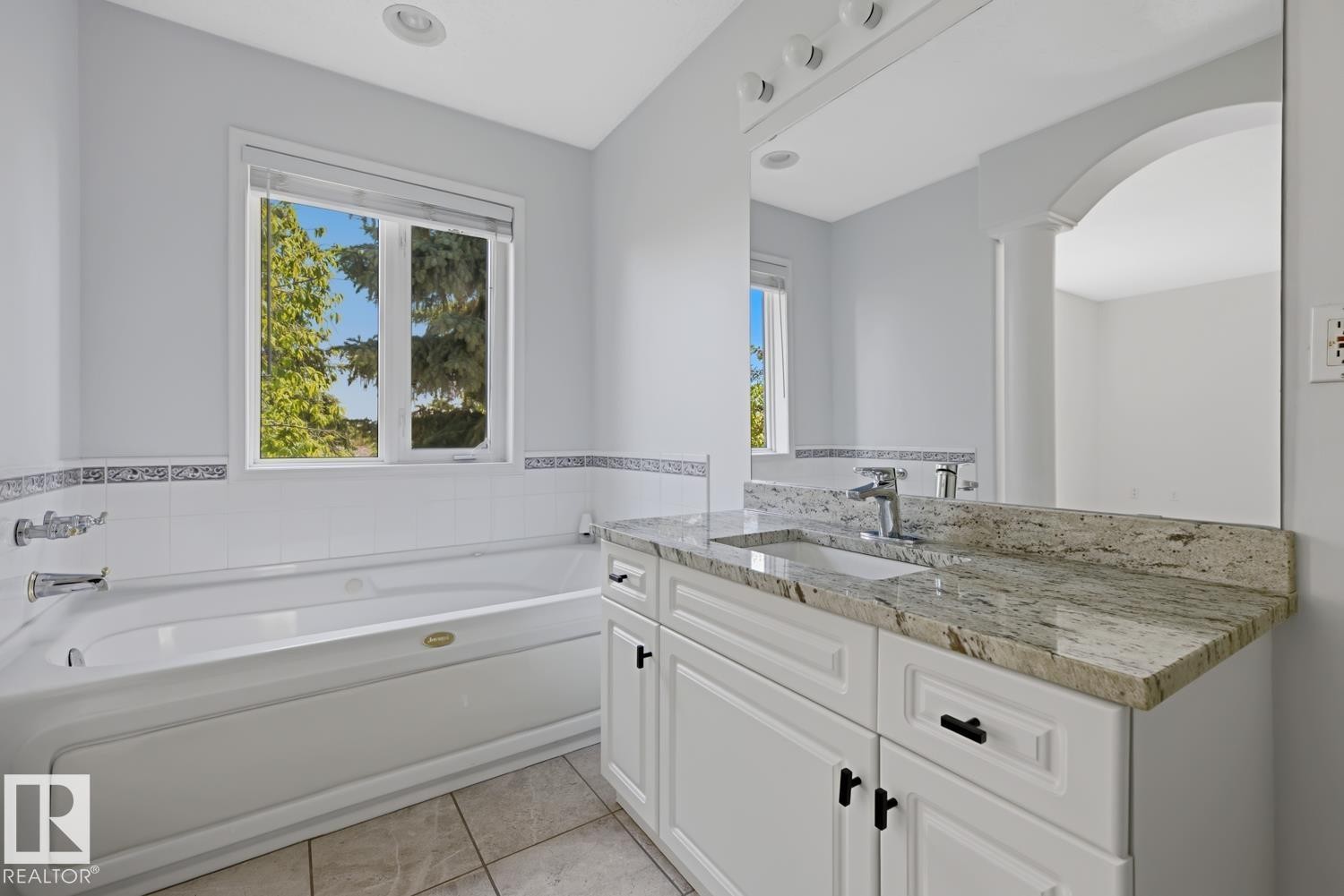 Bathroom featuring a jetted tub, a window with exterior scenery, a vanity with a granite countertop, an undermount sink, and white cabinetry with black hardware - 6 1237 Carter Crest Road, Edmonton, AB