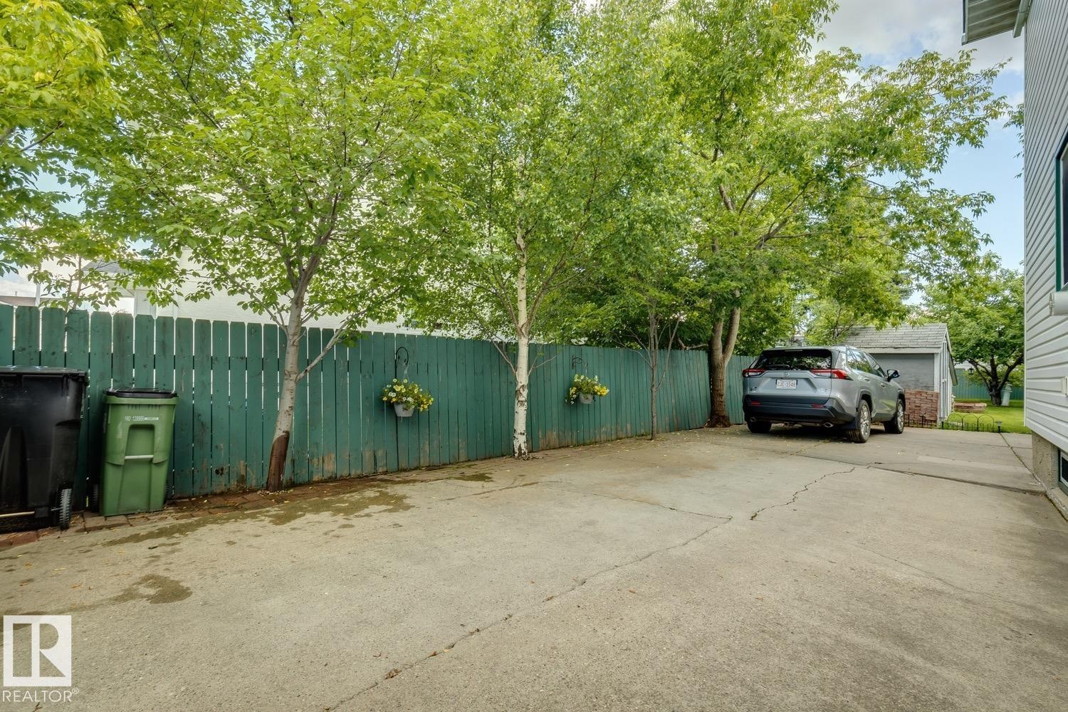 Paved driveway with a green wood fence, mature trees, and a detached structure in the background - 3015 131 Avenue, Edmonton, AB