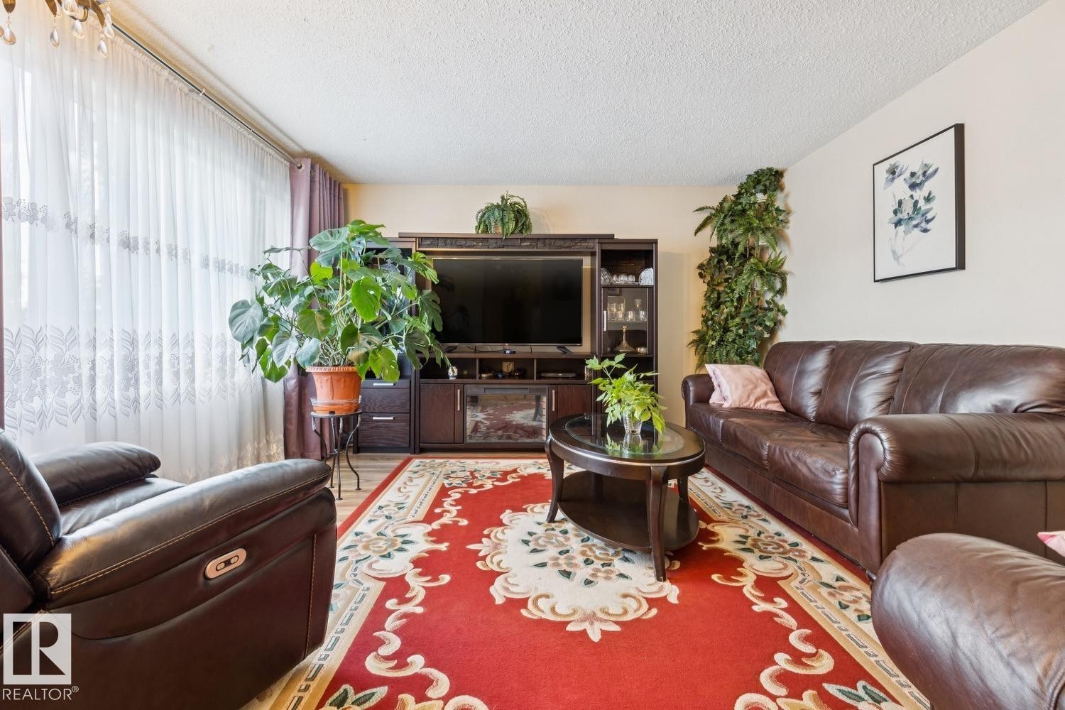 Spacious living area featuring wood-finish flooring, a decorative area rug, and a large window with sheer and solid drapes - 3015 131 Avenue, Edmonton, AB