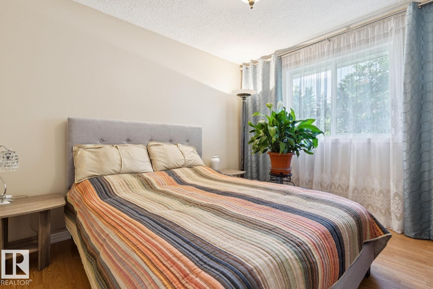 Bedroom featuring a large window with sheer and patterned drapes, wood-finish flooring, and light-toned walls - 3015 131 Avenue, Edmonton, AB