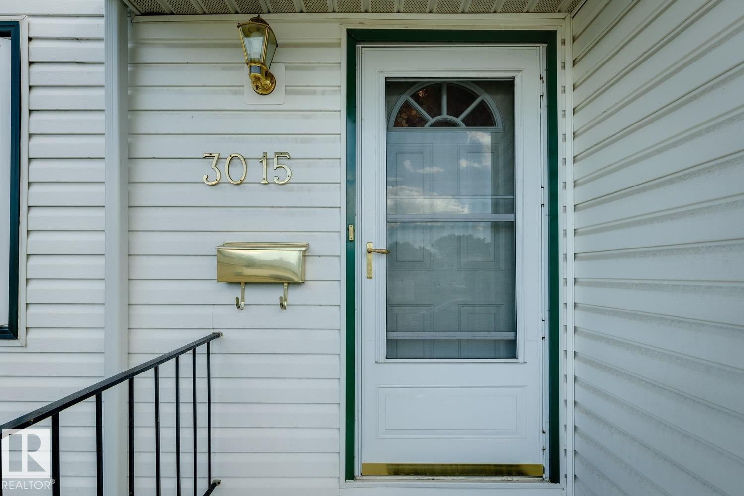 White siding exterior featuring a storm door with an arched window, brass house numbers, a coordinating brass mailbox, and a black metal handrail - 3015 131 Avenue, Edmonton, AB