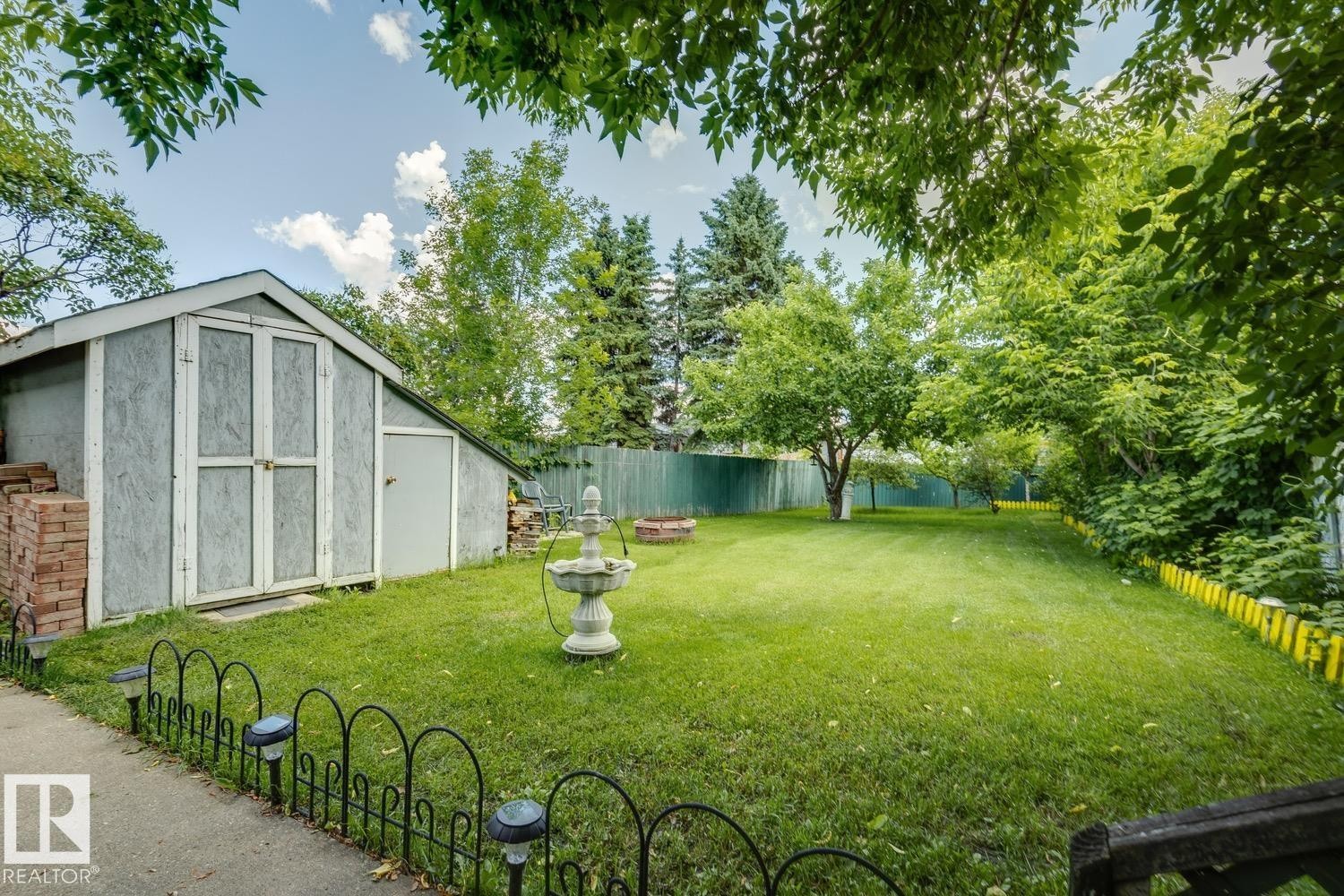 Spacious green lawn featuring a storage shed with double doors and a single side door, mature trees, a privacy fence, and decorative border fencing - 3015 131 Avenue, Edmonton, AB