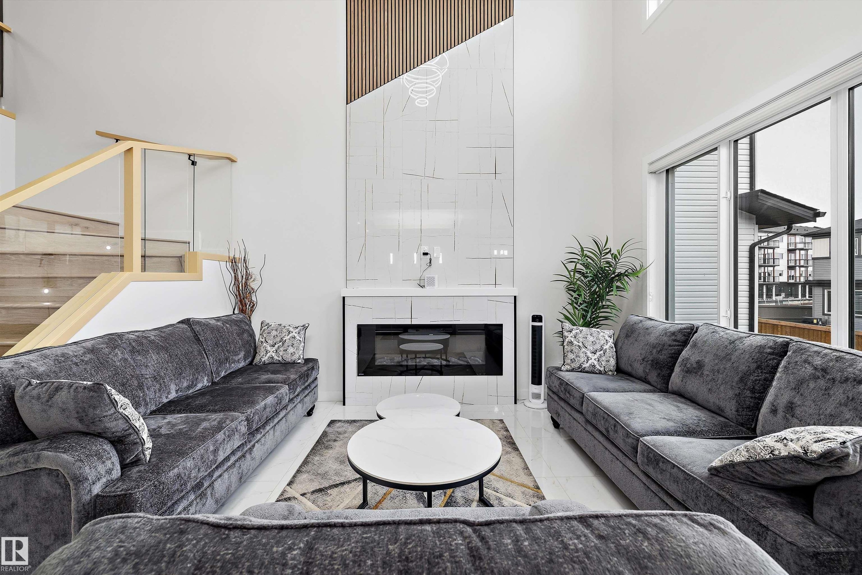 Two-story living area featuring a fireplace with a tile surround, tall windows, wood-finish flooring, and a staircase with a glass railing - 403 Pine Point, Leduc, AB
