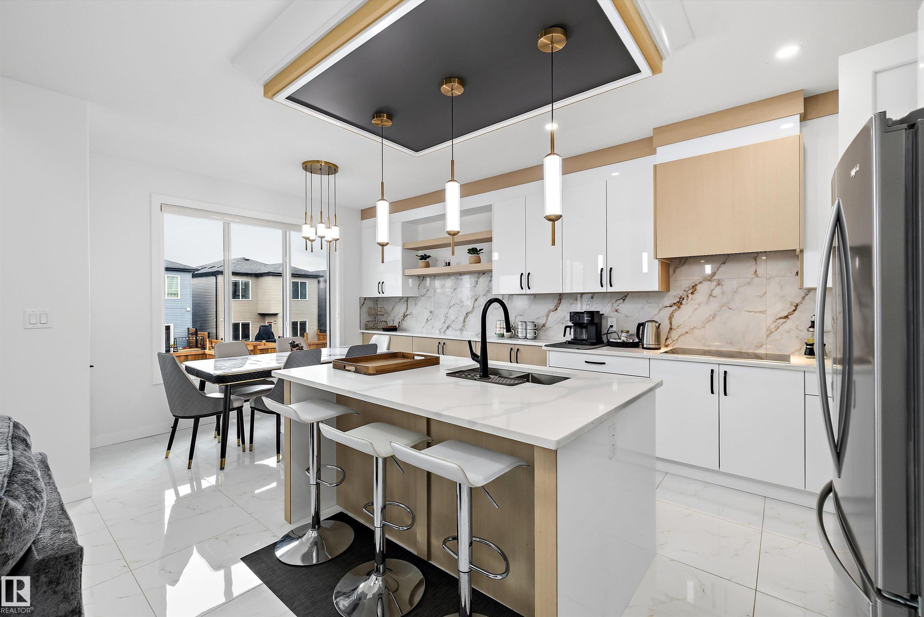 Gourmet kitchen featuring a central island with countertop seating, high-gloss white cabinetry, gold-tone accents, and a full-height marble-look backsplash - 403 Pine Point, Leduc, AB