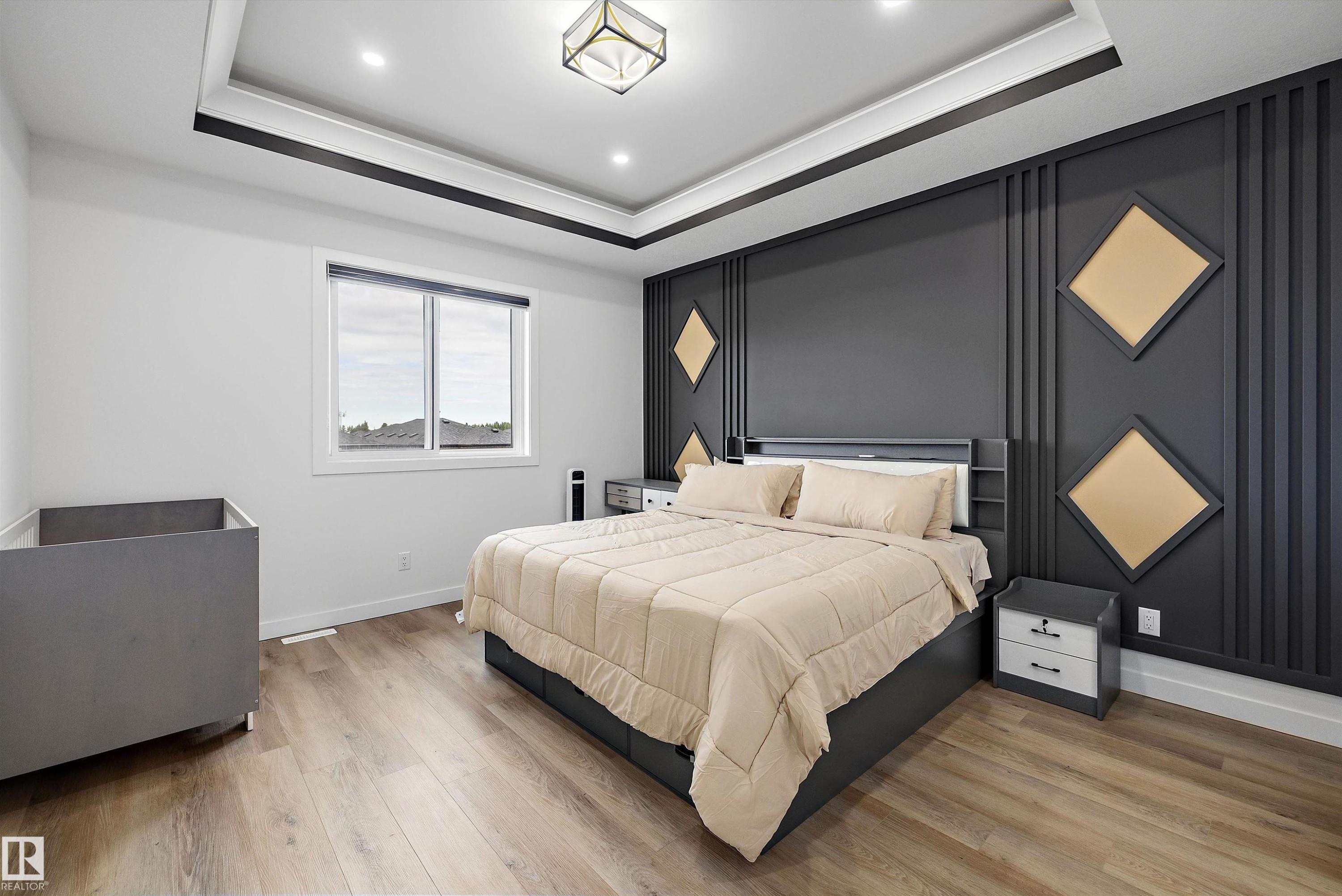 Bedroom featuring a decorative accent wall with geometric paneling, tray ceiling with recessed lighting, central flush-mount fixture, wood-finish flooring, and a large window - 403 Pine Point, Leduc, AB
