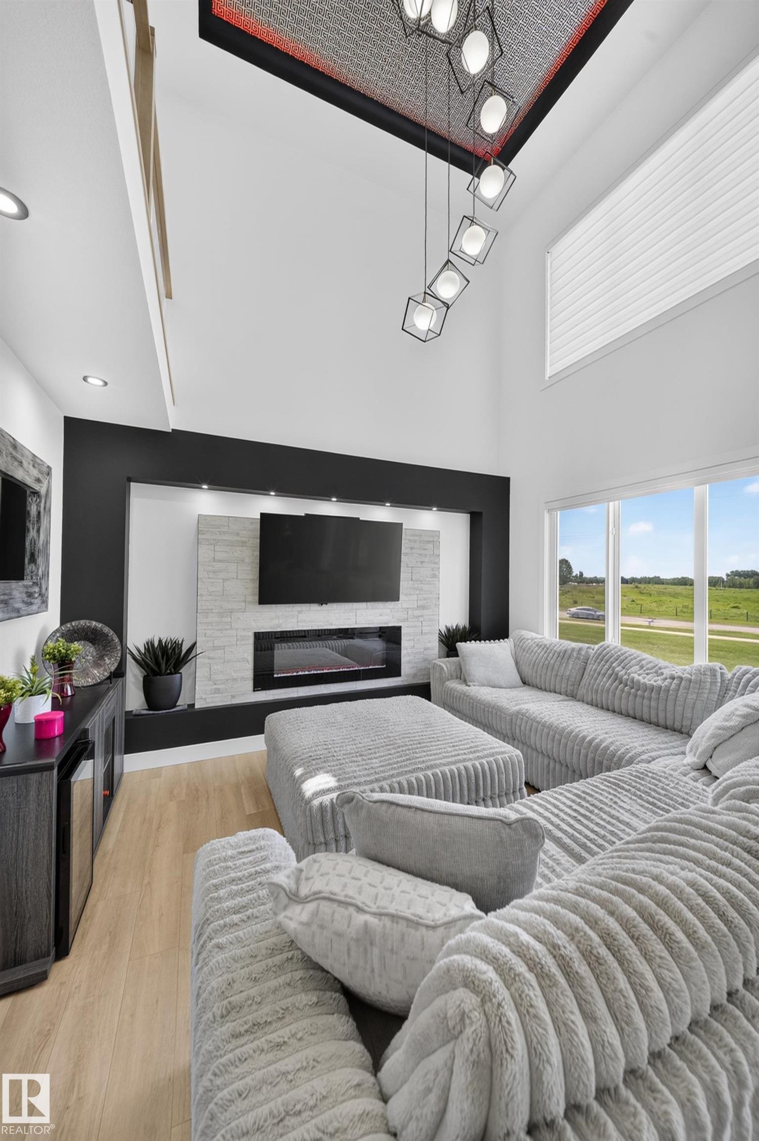 Vaulted ceiling with a geometric chandelier, integrated entertainment wall with an electric fireplace, wood-finish flooring, and large windows - 626 Astoria Way, Devon, AB