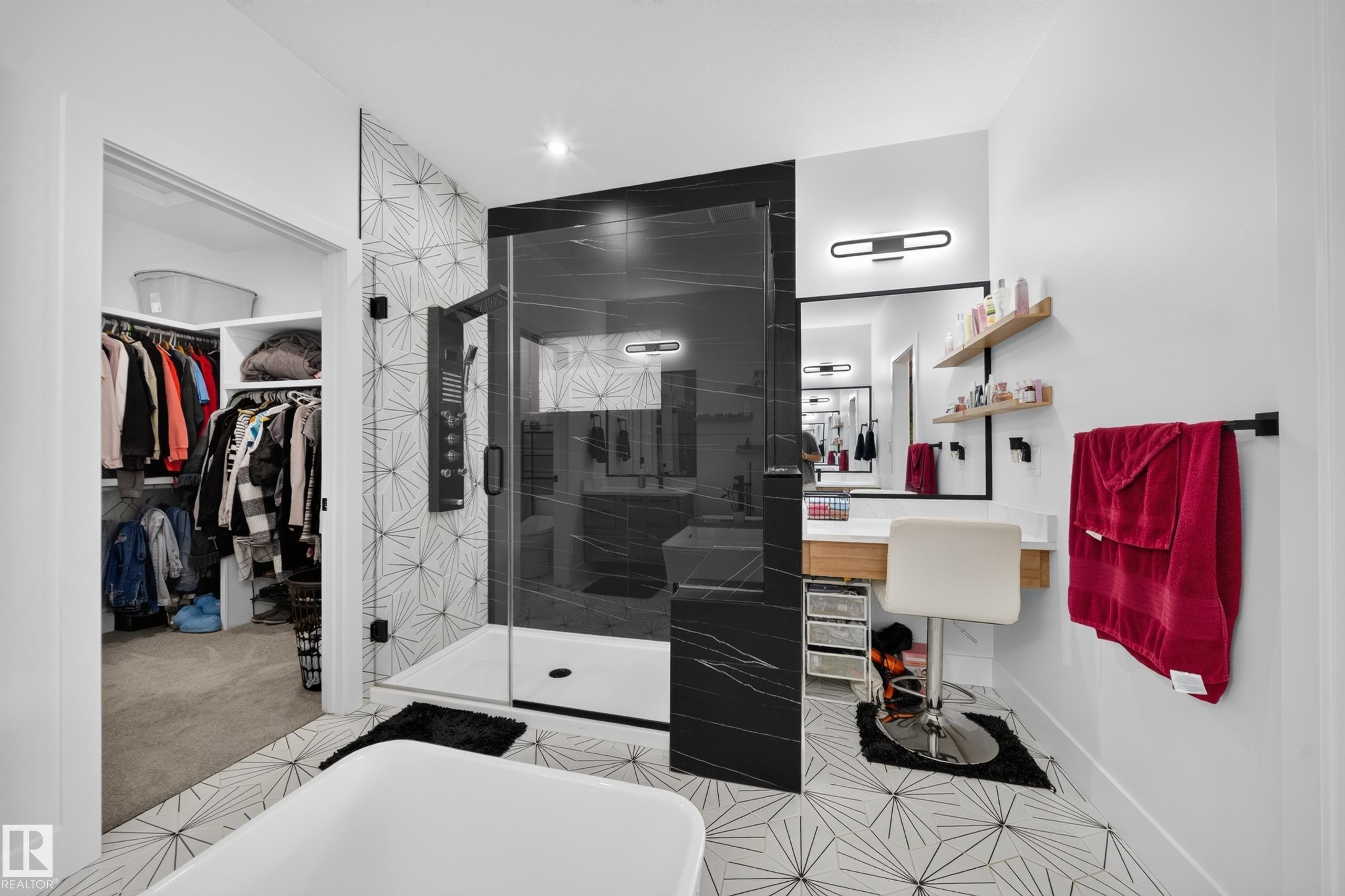 Modern bathroom featuring geometric tile flooring, a glass-enclosed shower with black marble-look paneling and a multi-function shower system, a built-in vanity area with floating shelves, and an open closet system - 626 Astoria Way, Devon, AB