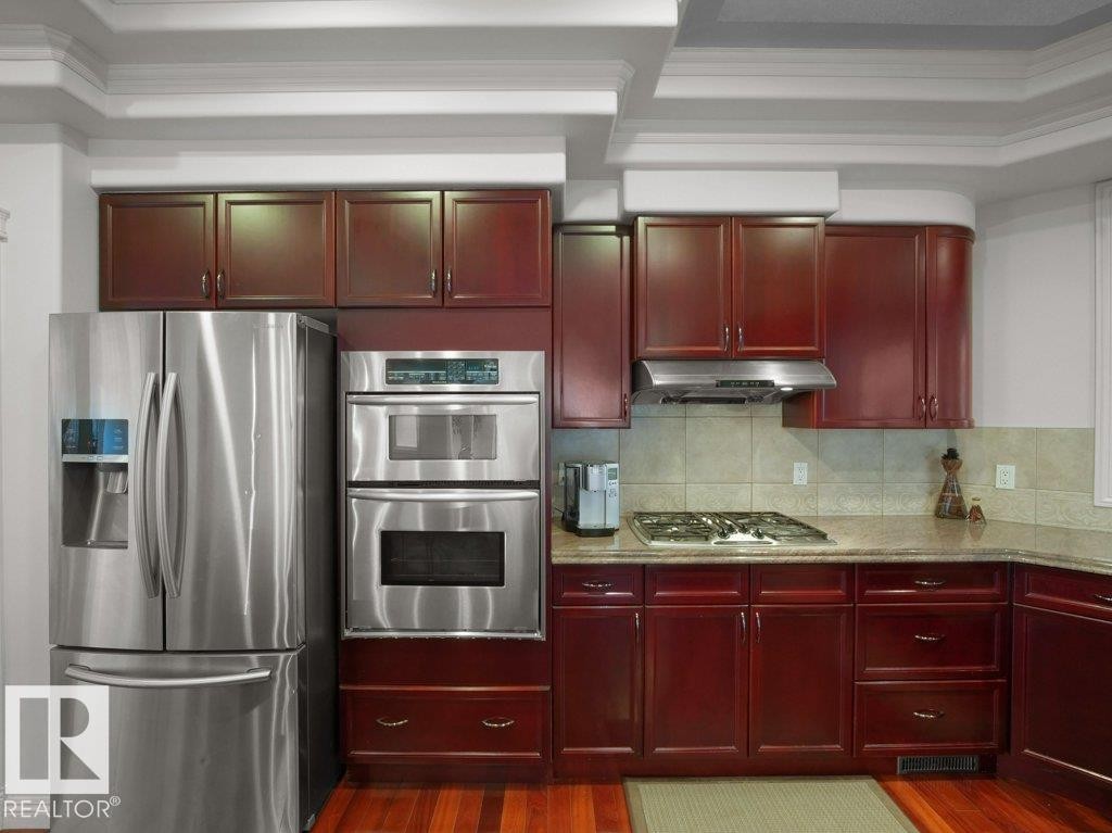 Kitchen featuring rich wood cabinetry, stainless steel appliances including a refrigerator and double wall ovens, a gas cooktop with a range hood, and light-toned countertops - 2423 Tegler Green, Edmonton, AB
