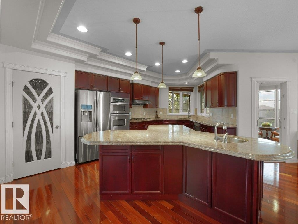Kitchen featuring rich wood cabinetry, stainless steel appliances, a curved island with a stone countertop, and pendant lighting - 2423 Tegler Green, Edmonton, AB
