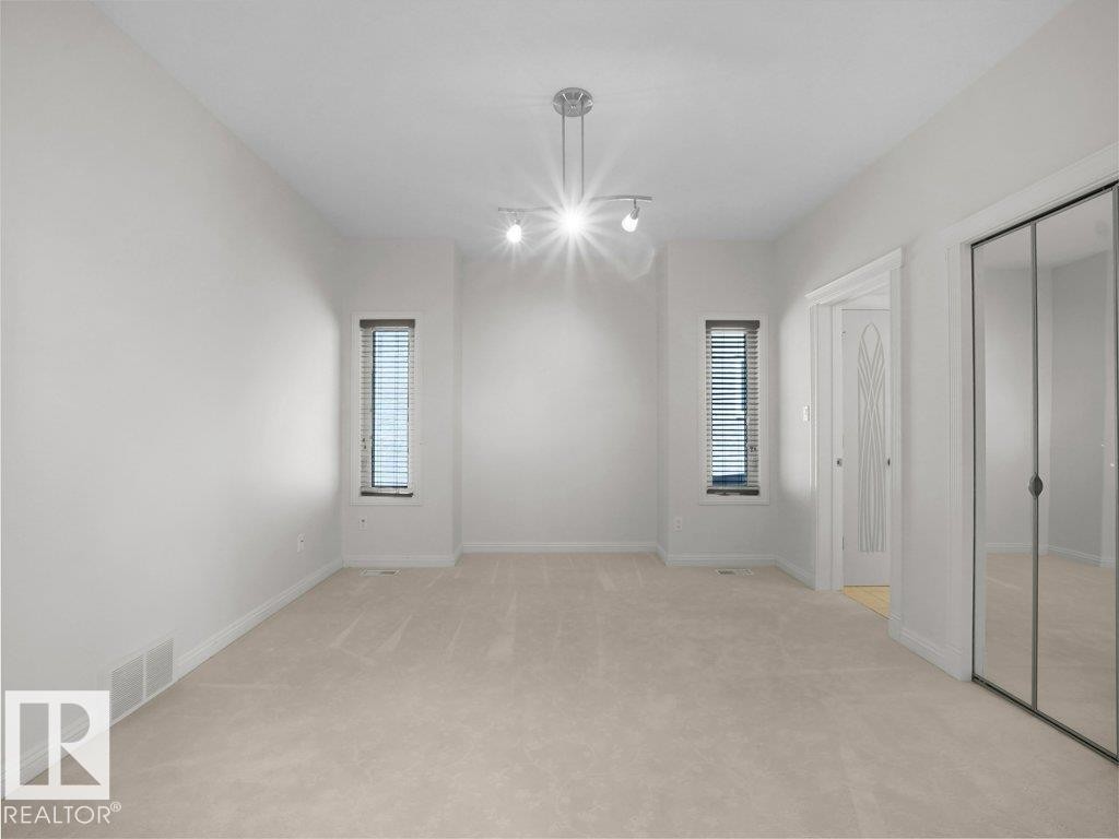 Spacious room featuring neutral carpeting, white walls, two vertical windows with blinds, a modern track light fixture, and mirrored closet doors - 2423 Tegler Green, Edmonton, AB
