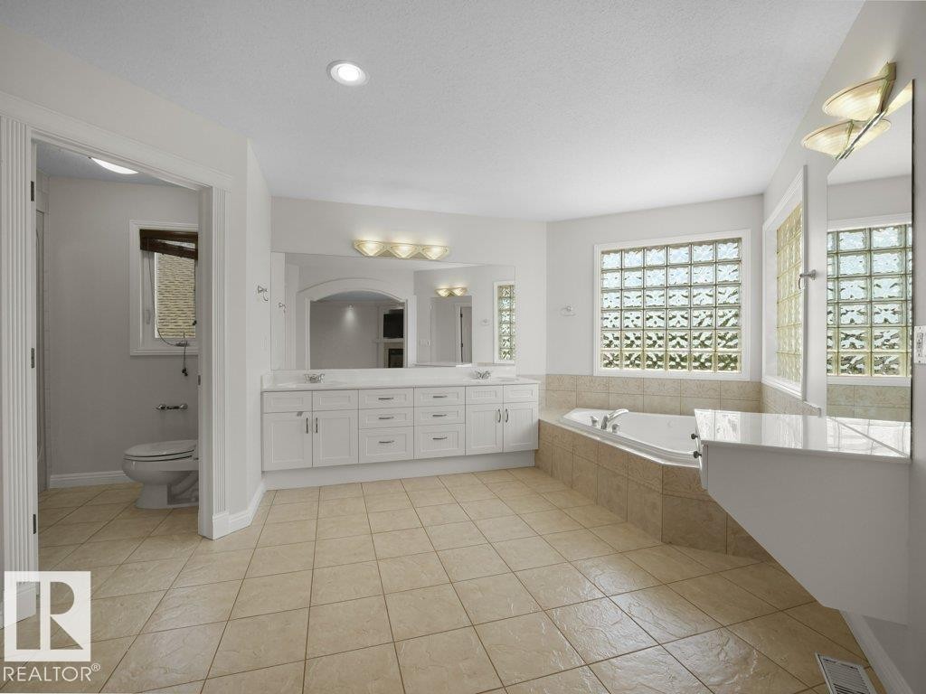 Spacious bathroom featuring a dual vanity with white cabinetry and an arched mirror, a built-in soaking tub with tiled surround, and glass block windows - 2423 Tegler Green, Edmonton, AB