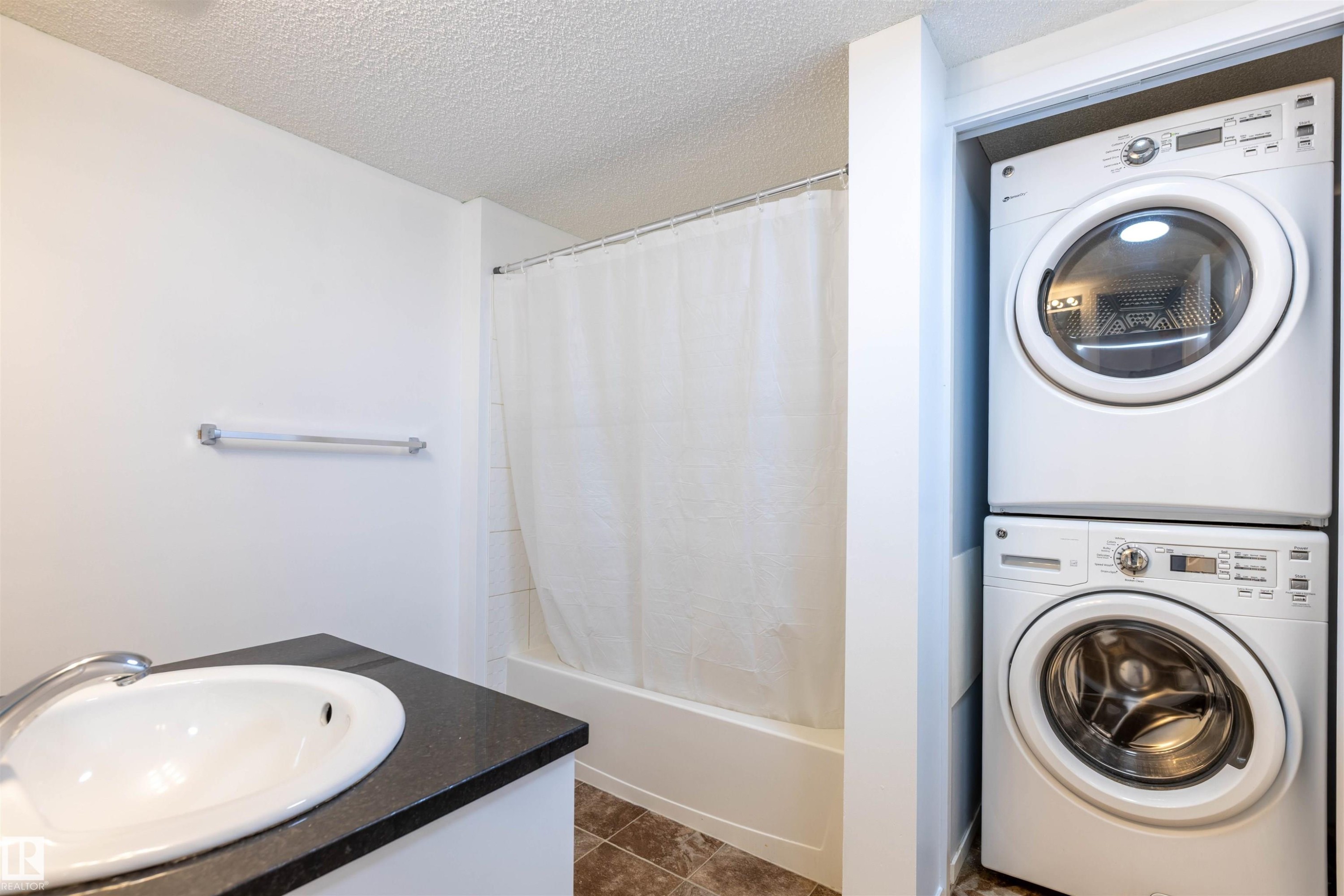 Bathroom featuring a single vanity with a white basin and dark countertop, a tub and shower combination with a white shower curtain, and brown tiled flooring - 403 25 Element Dr, St. Albert, AB - Indoor Photo Showing Laundry Room
