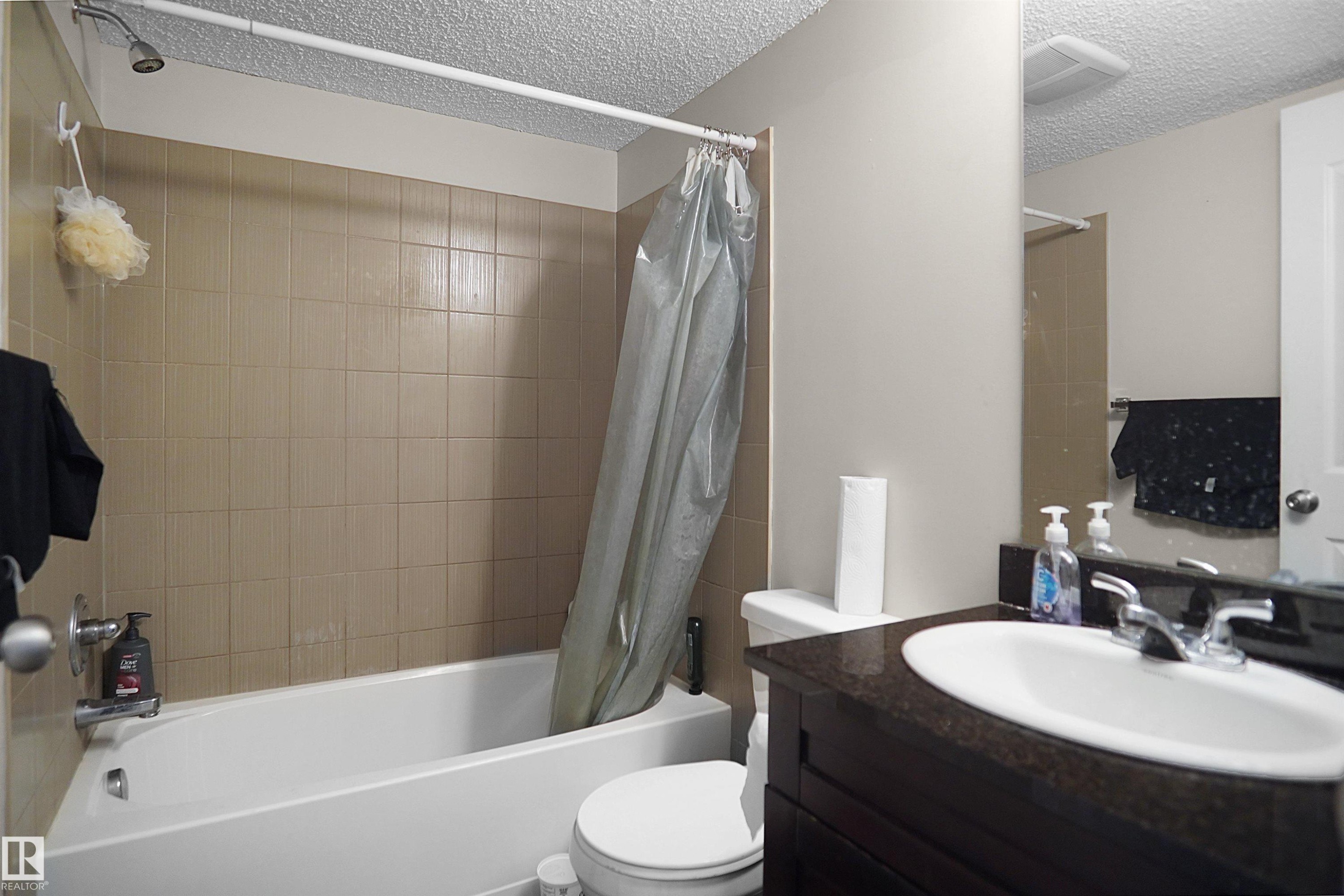 Bathroom featuring a white porcelain tub, tan ceramic tile surround, dark wood-finish vanity with an undermount sink, and a dark stone-finish countertop - 215 1070 Mcconachie Blvd, Edmonton, AB