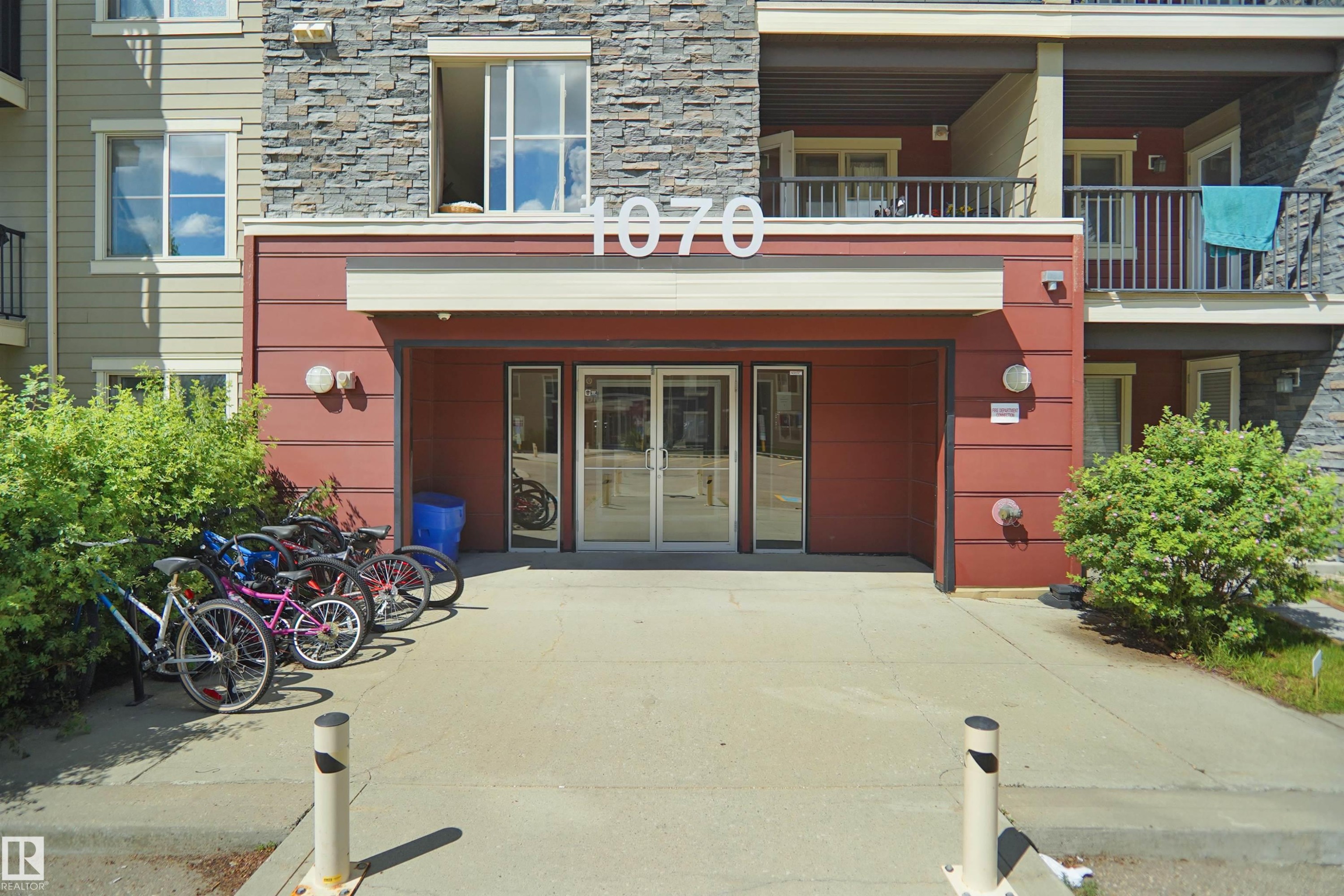 Building entrance featuring double glass doors, a stone facade, and red paneling - 215 1070 Mcconachie Blvd, Edmonton, AB