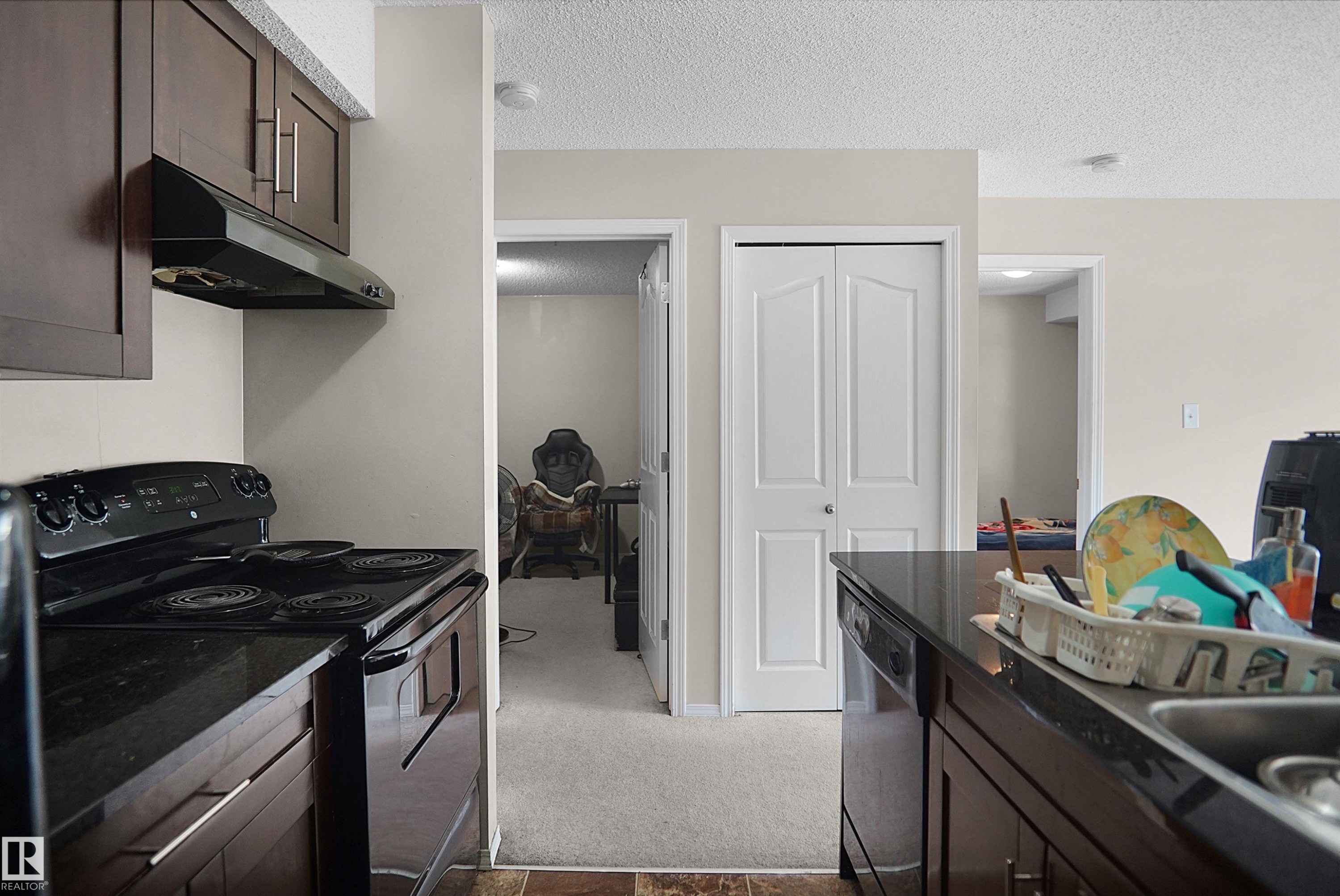 Kitchen featuring dark wood-finish cabinetry, black electric range with coordinating range hood, and dark countertops - 215 1070 Mcconachie Blvd, Edmonton, AB