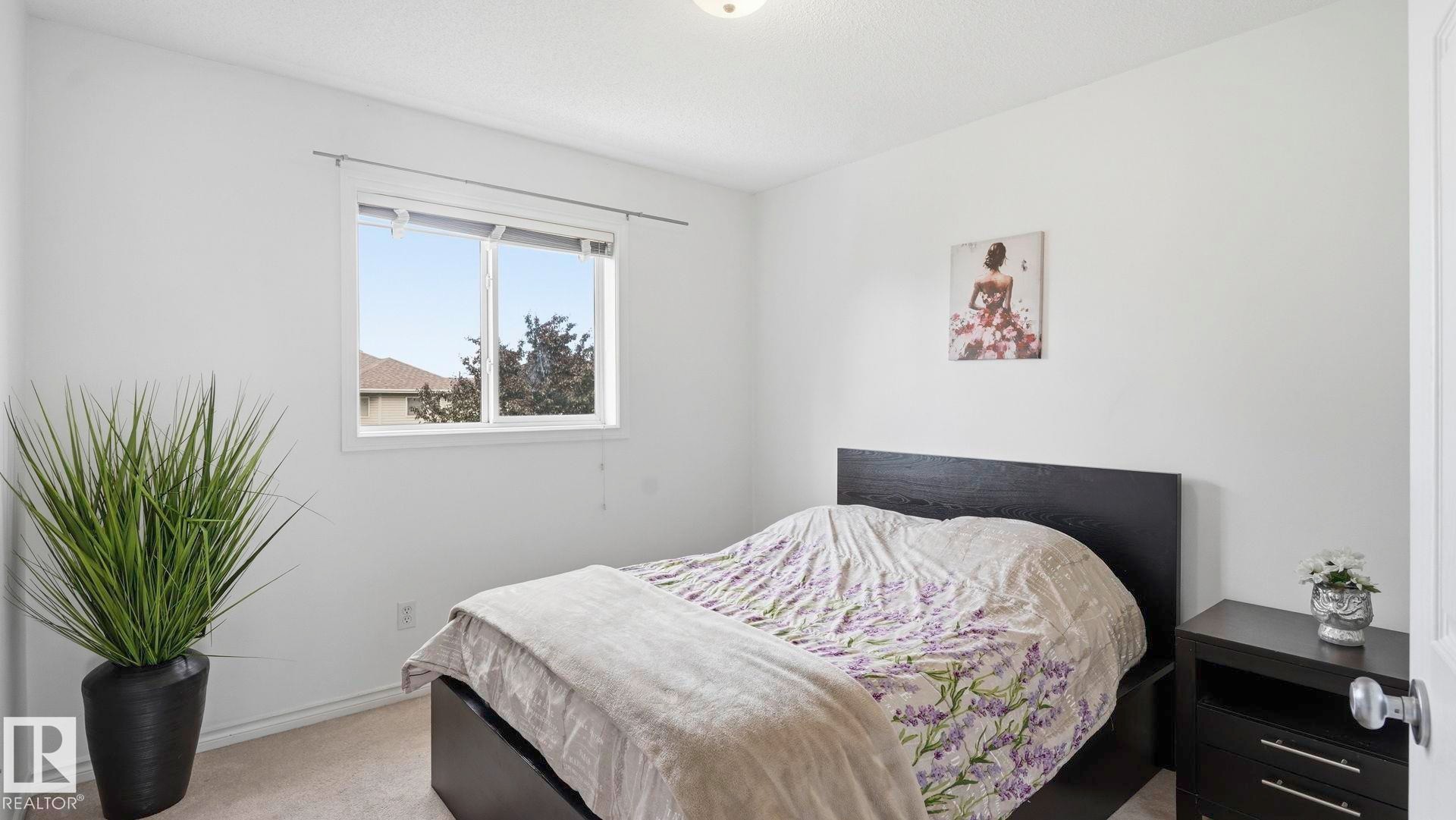 Bright room featuring a window with white trim, light-colored carpet, white walls, a dark headboard, and a dark bedside table - 3591 Mclean Crescent, Edmonton, AB