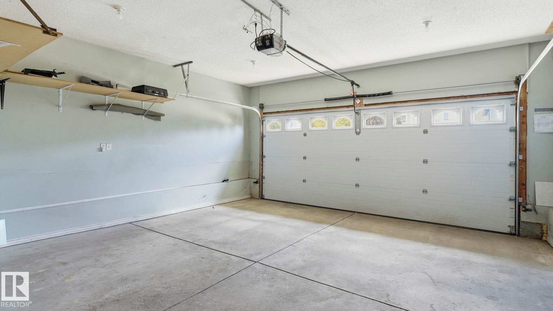 Spacious garage featuring a concrete floor, overhead lighting, an automatic garage door opener, and built-in wall shelving - 3591 Mclean Crescent, Edmonton, AB