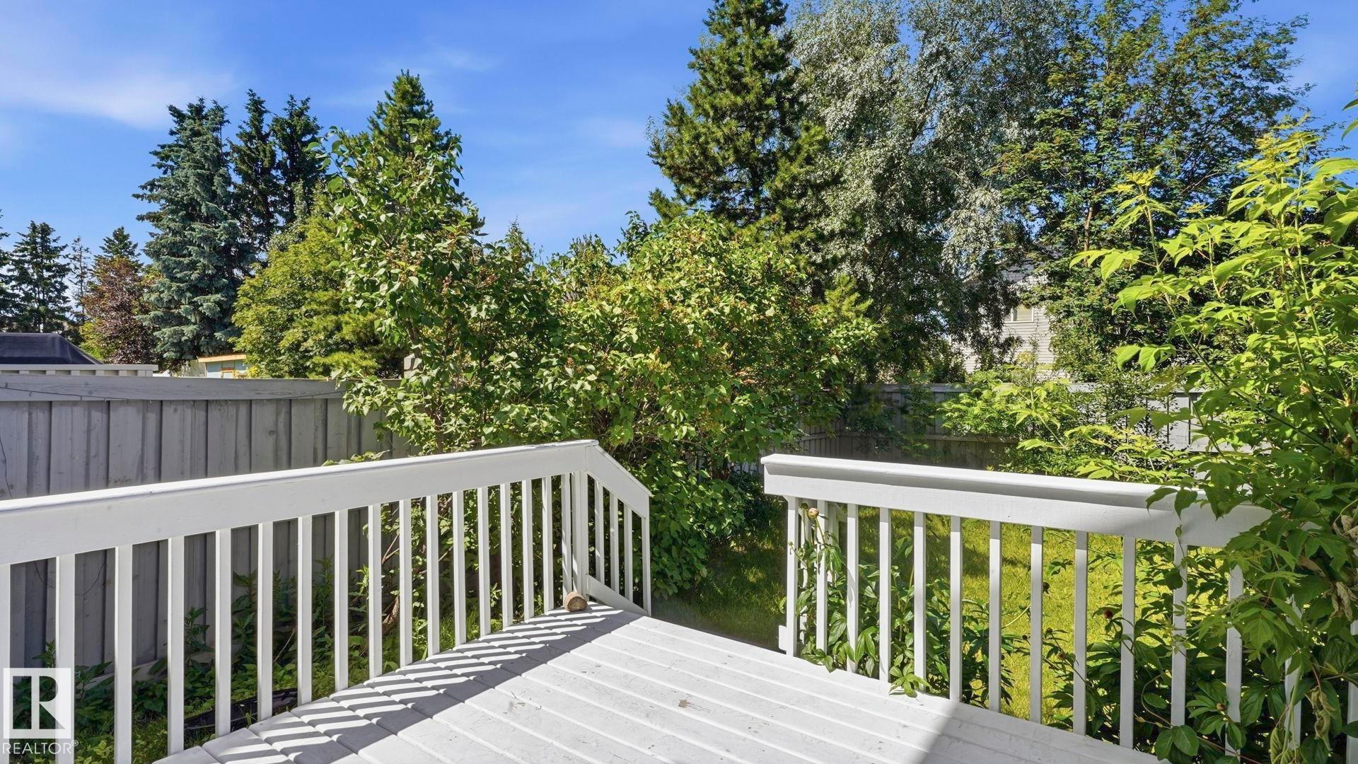 White wood deck featuring a baluster railing, surrounded by mature trees, shrubs, and a privacy fence - 3591 Mclean Crescent, Edmonton, AB
